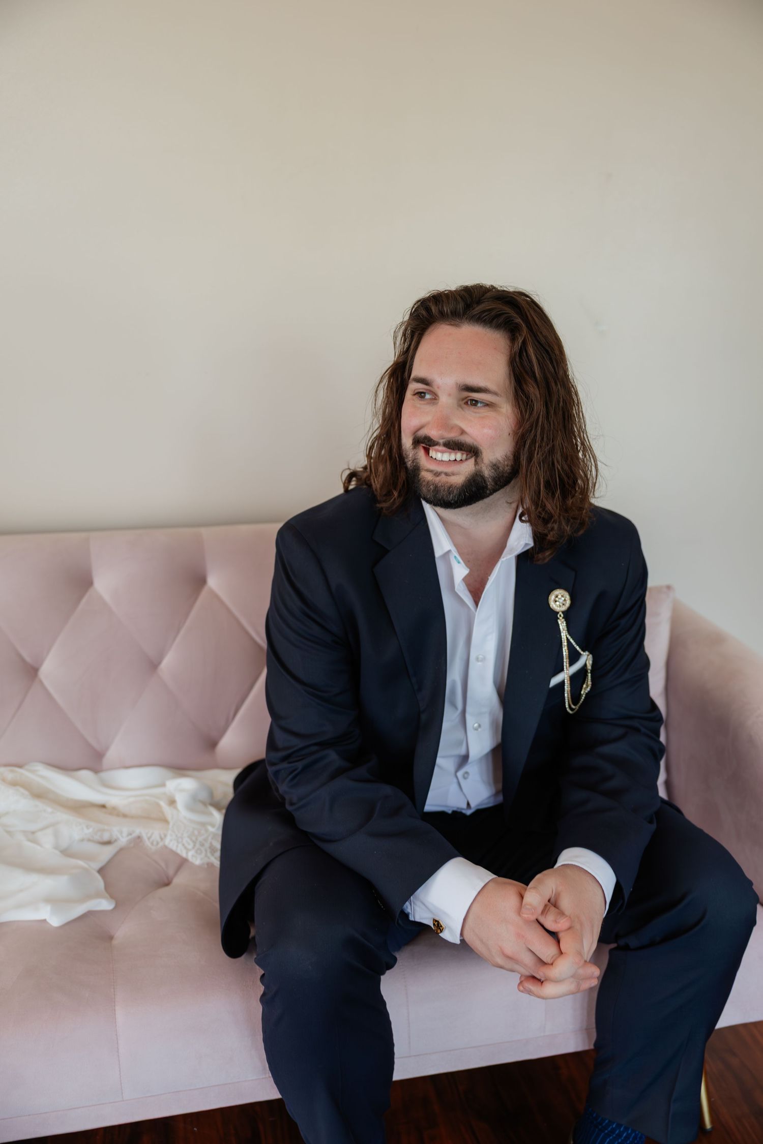 Smiling groom with long hair and beard in navy suit with bolo tie seated on pink velvet sofa.