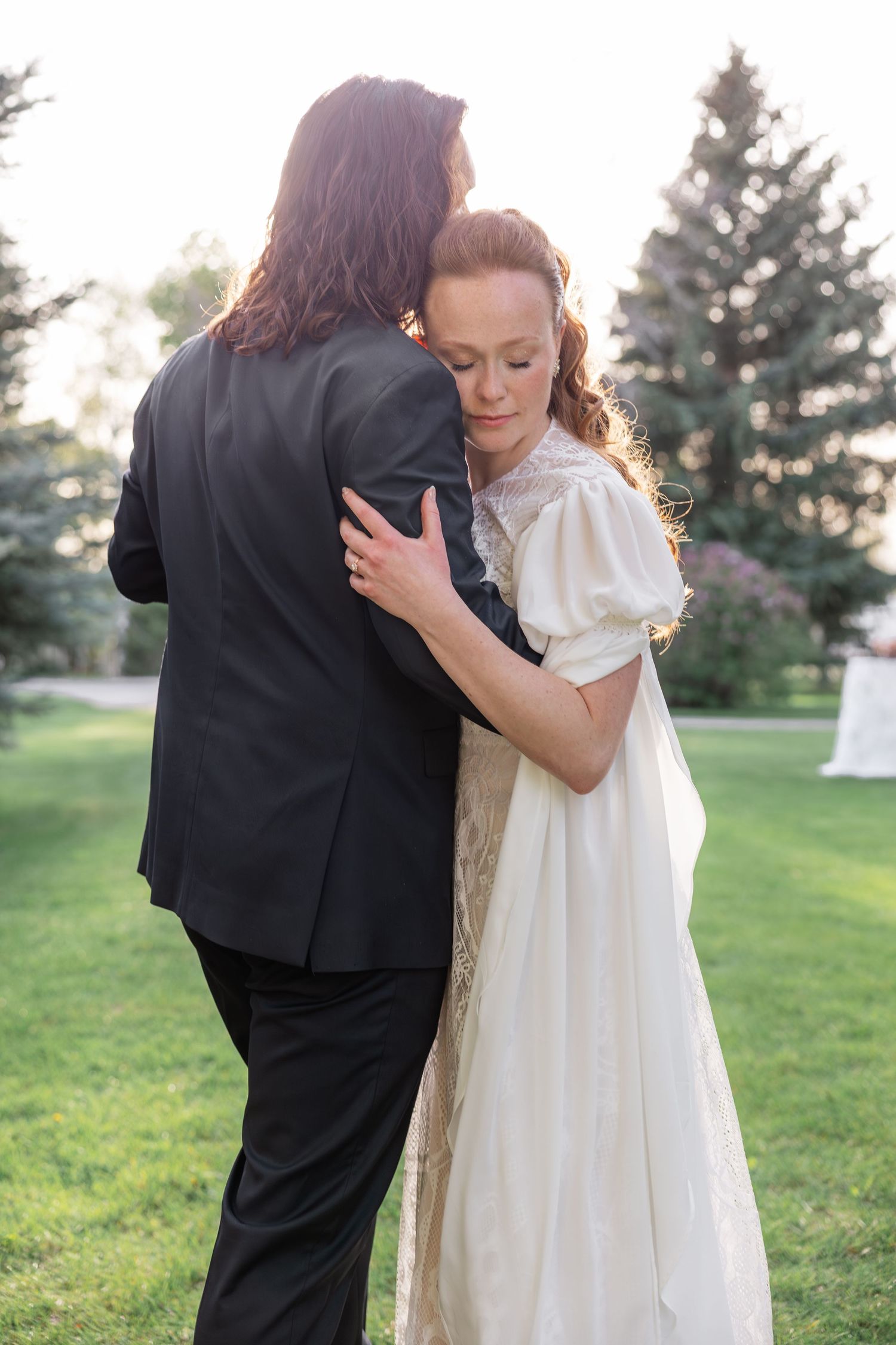 Bride in vintage white gown embraces groom outdoors at golden hour in lush green garden.