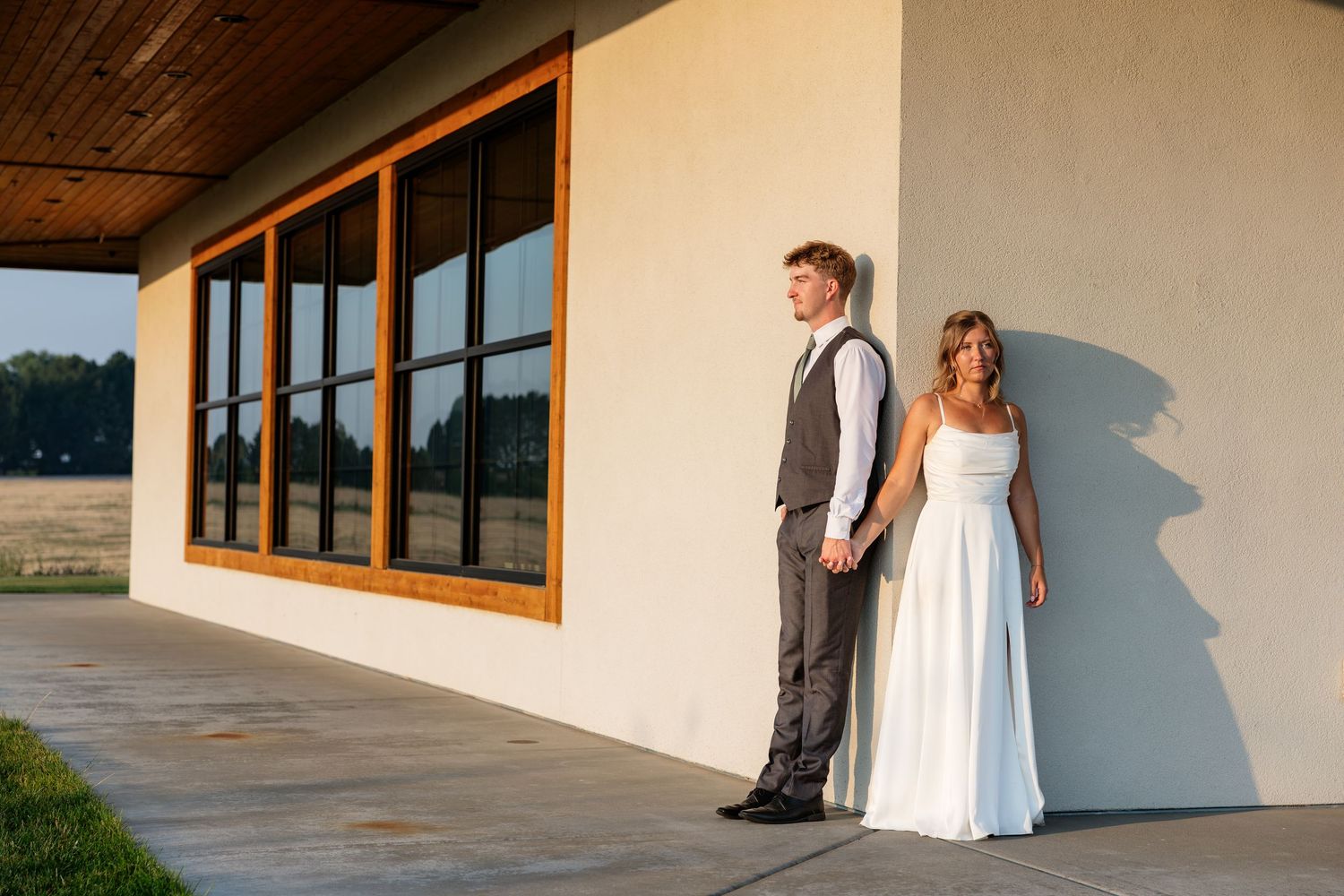 Bride and groom hold hands against a white barn wall at golden hour during their rustic outdoor wedding.