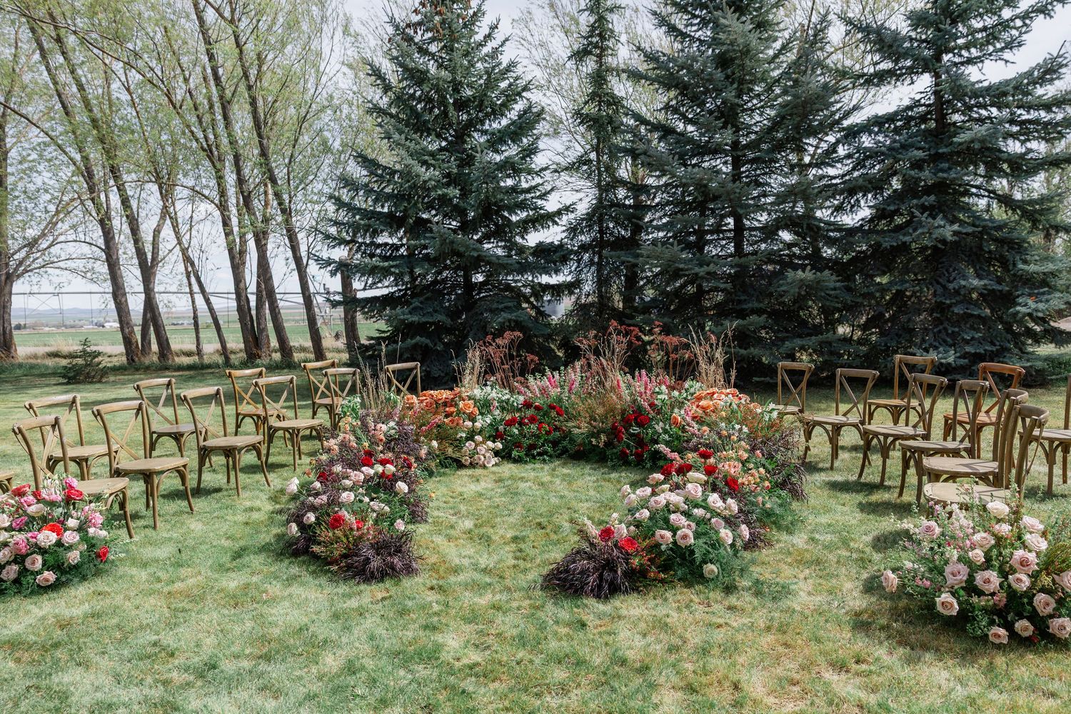 Outdoor wedding ceremony setup with floral arrangements in a circular pattern on green grass surrounded by tall evergreen trees.