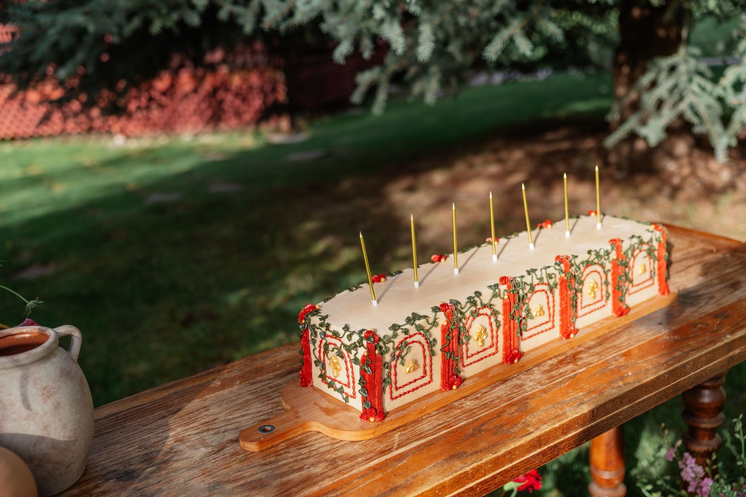 Rectangular white wedding cake decorated with red floral details and gold candles on a rustic wooden table.