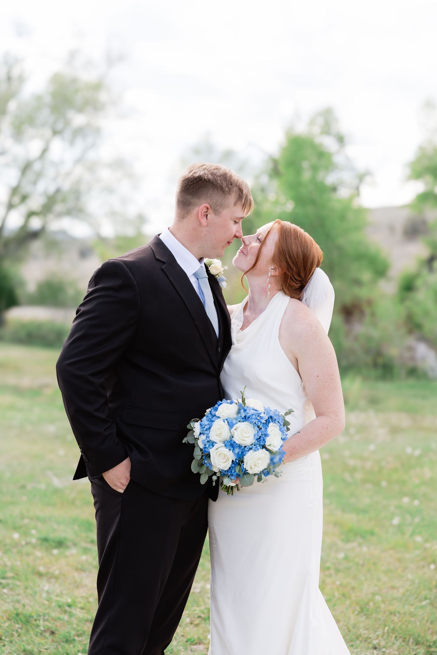 Bride and groom share a romantic moment outdoors, she holds a blue and white bouquet, wearing a white gown and veil.