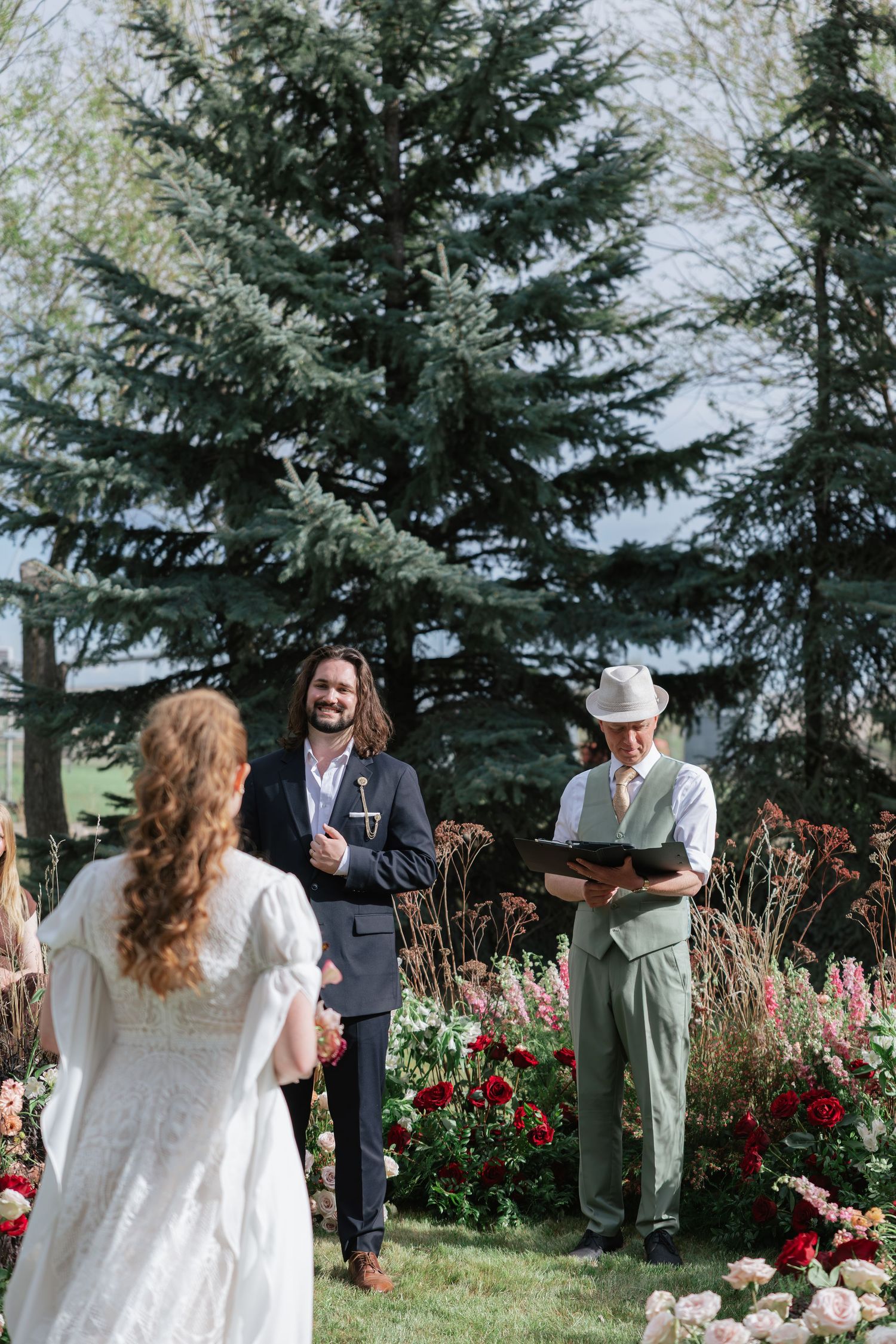 Outdoor wedding ceremony with groom in dark suit, officiant in white hat, bride approaching floral altar with evergreen trees.