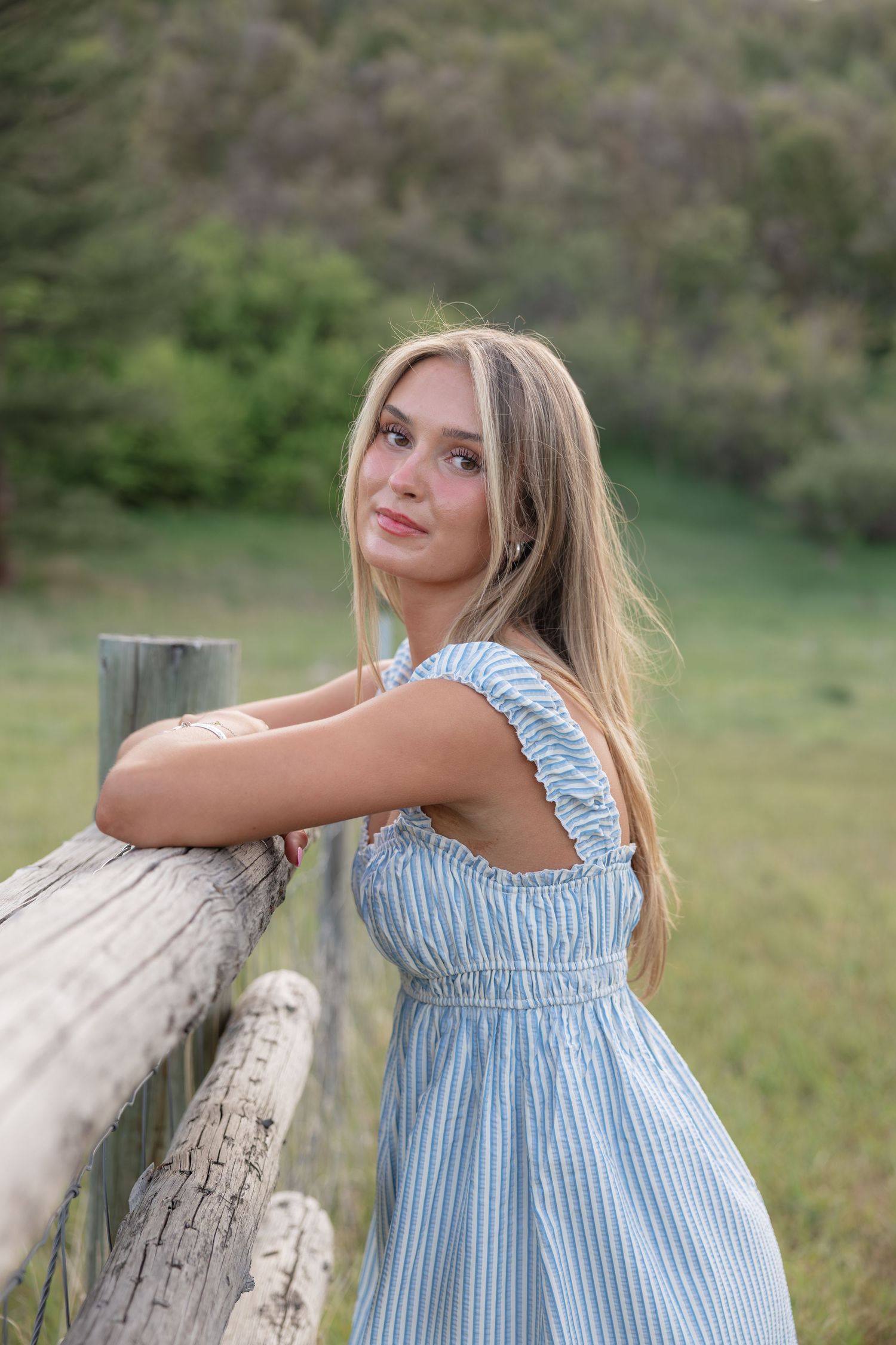 Young blonde woman in blue striped sundress leaning on rustic wooden fence in green countryside setting.