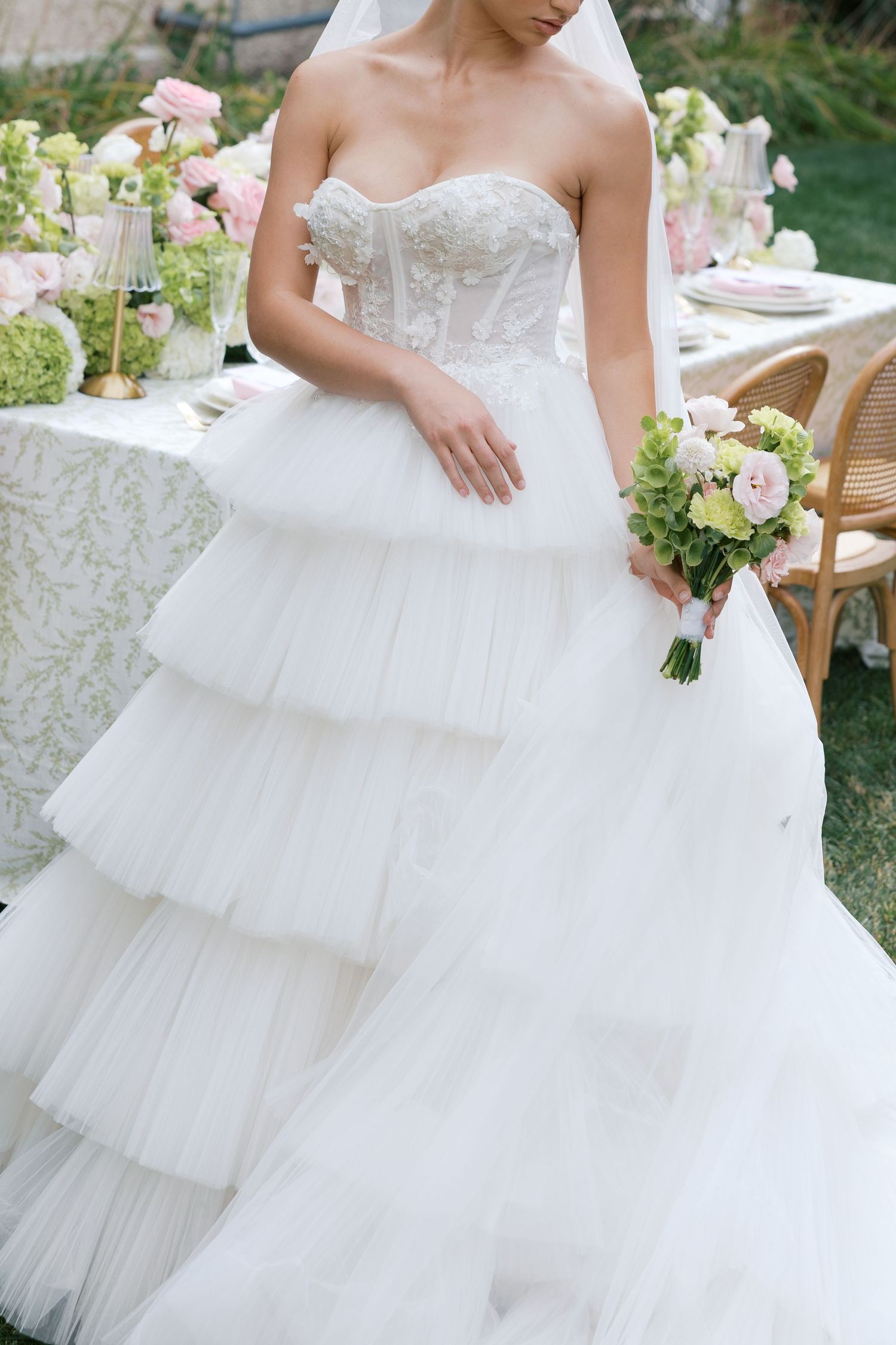 Bride in strapless tiered tulle ball gown with lace corset bodice holding pink and green bouquet at outdoor wedding reception.
