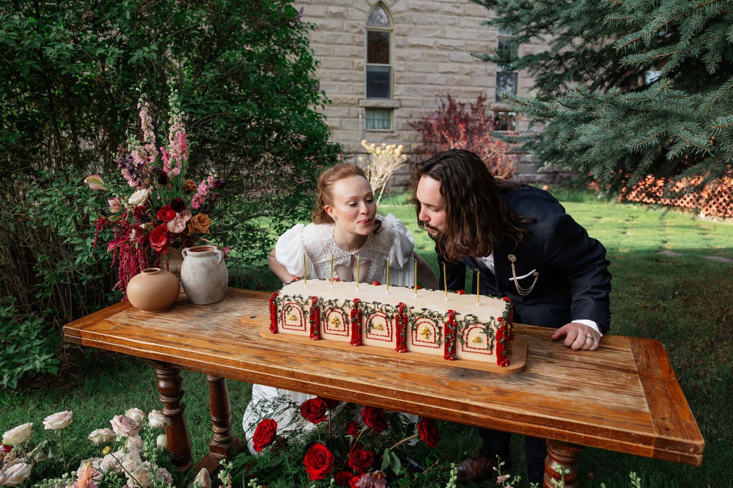 Bride and groom blow out candles on decorative cake display at outdoor rustic wedding reception.