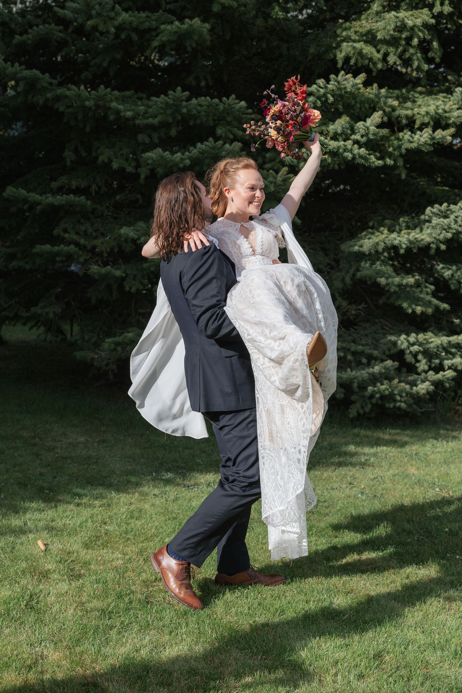 Joyful groom lifting his smiling bride in a lace wedding dress outdoors with evergreen trees behind them.