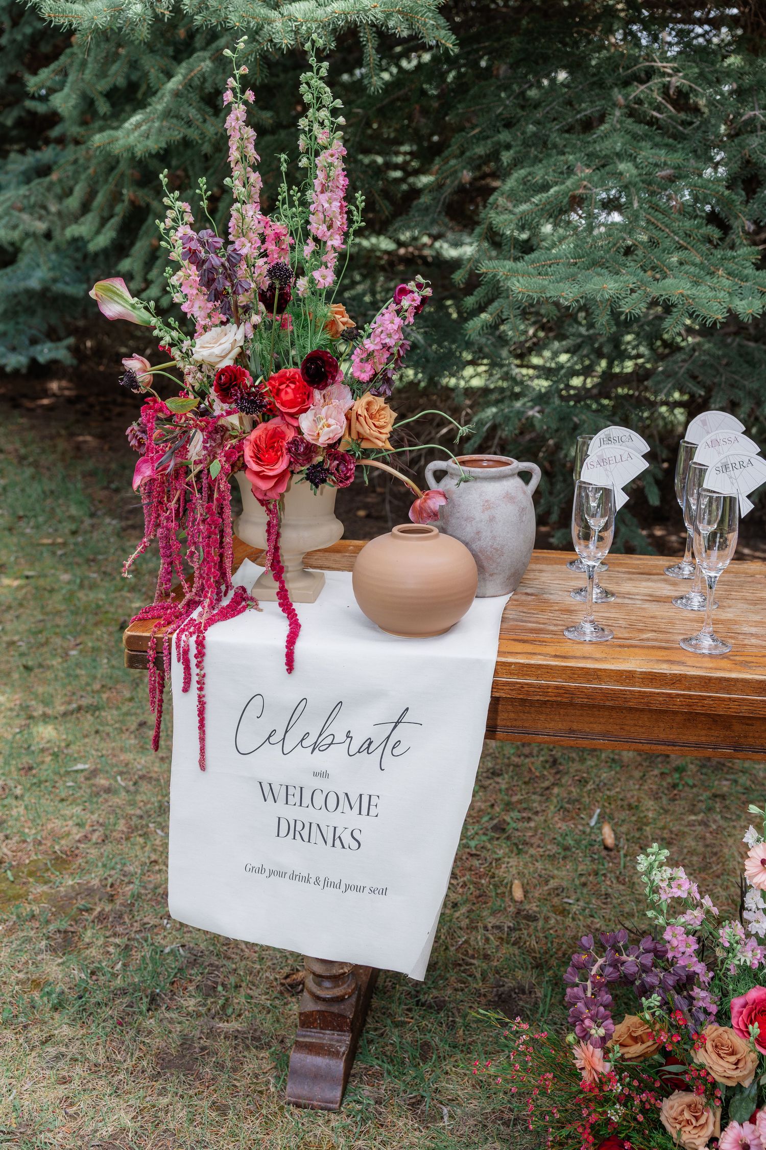 Elegant wedding welcome drinks table with vibrant floral arrangement, terracotta vases, and champagne flutes on a wooden bench outdoors.