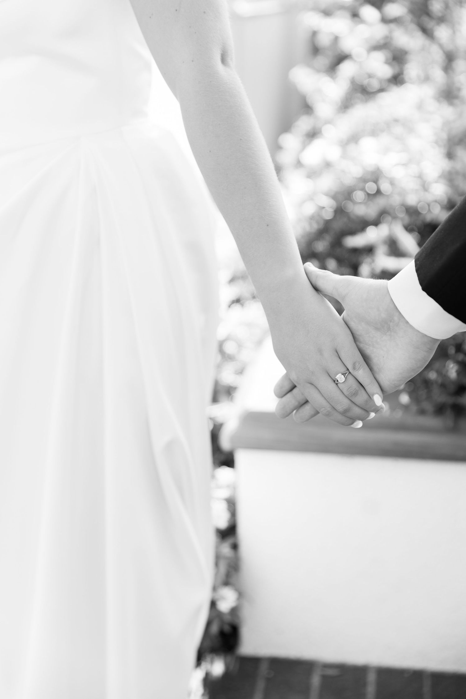 Black and white photo of bride and groom holding hands, showcasing wedding ring during outdoor ceremony.