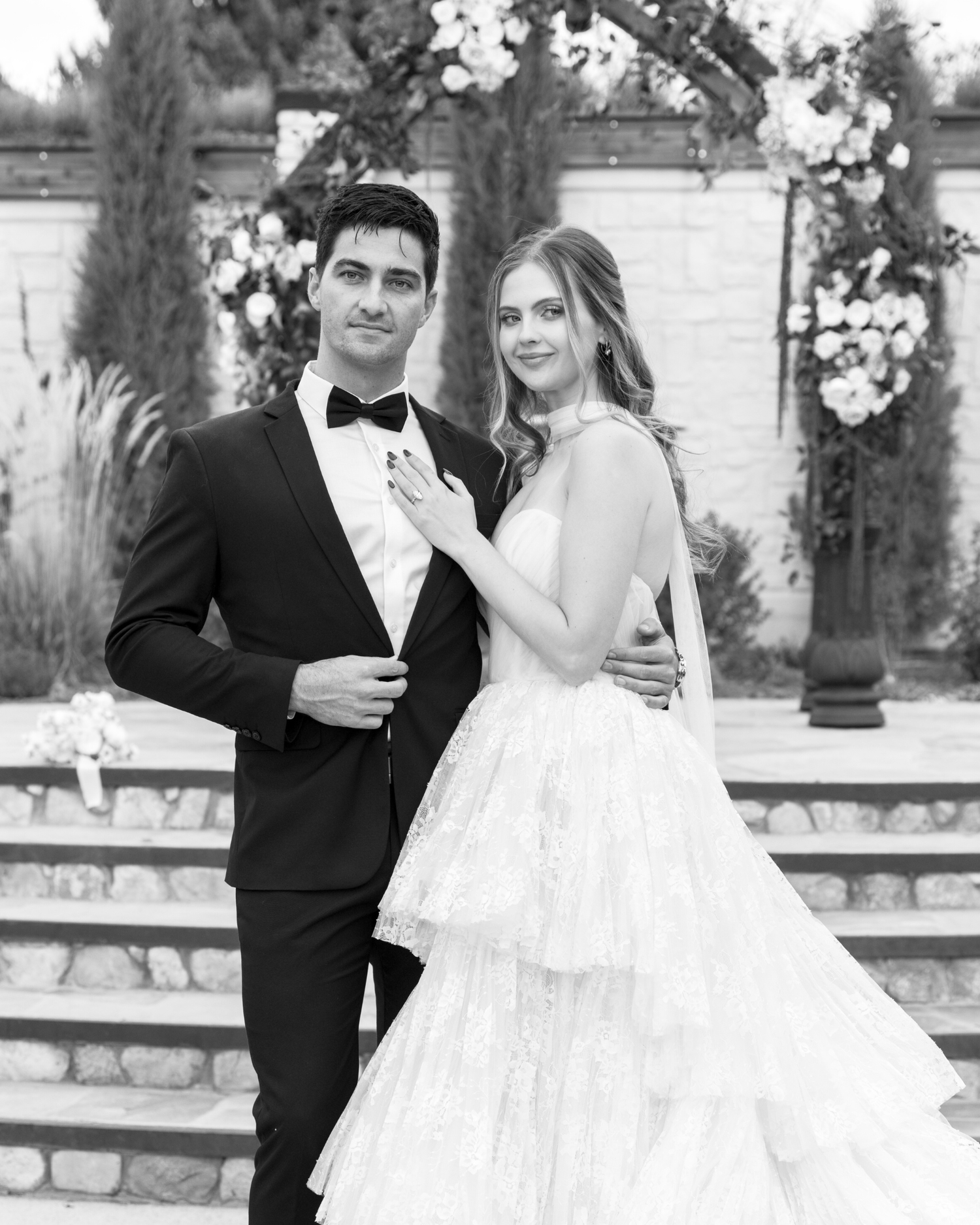 A couple in formal wedding attire poses together in a garden setting with stone architecture in the background.