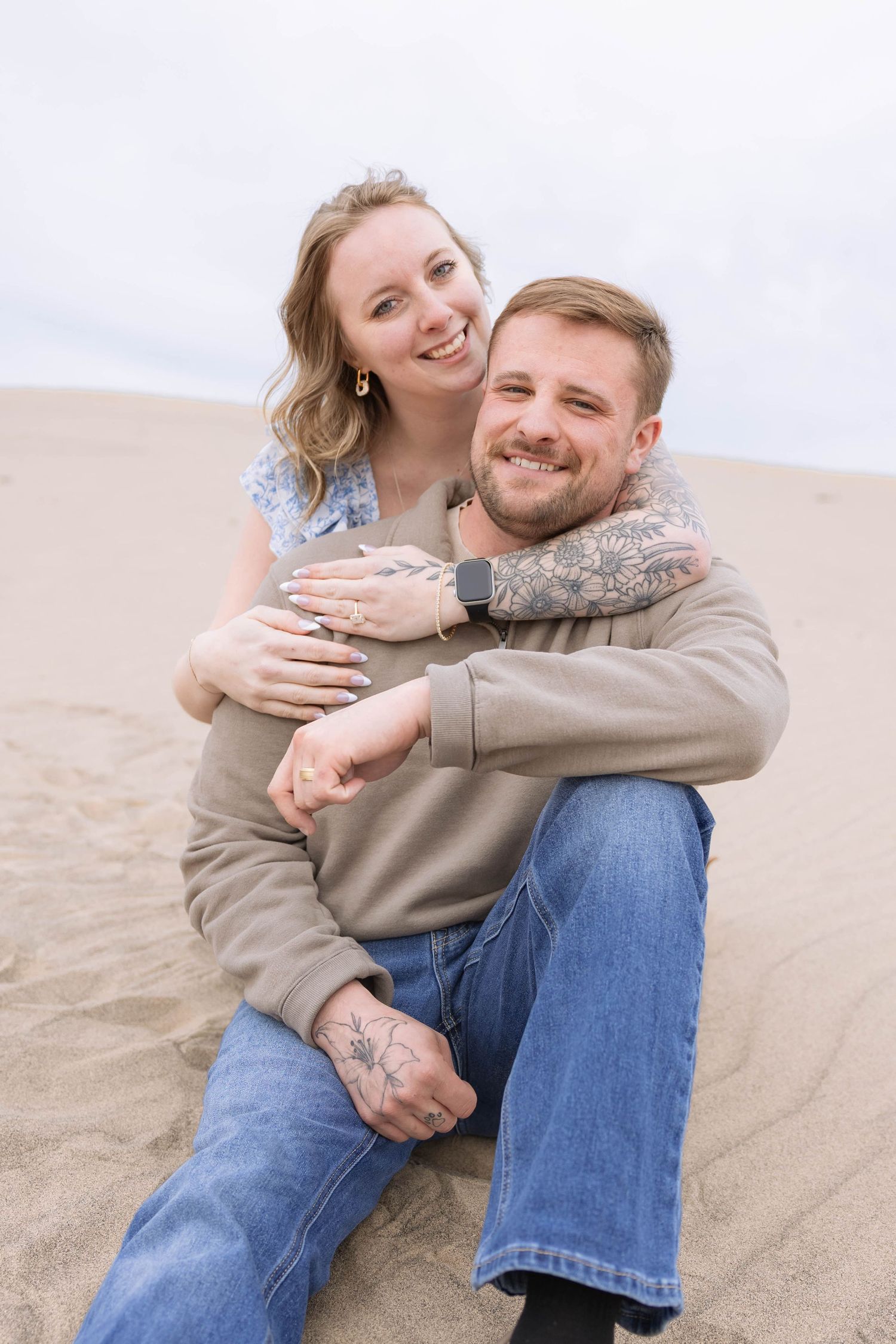 Happy couple embracing on sandy dunes, woman hugging man from behind, both smiling, wearing casual jeans and tops outdoors.