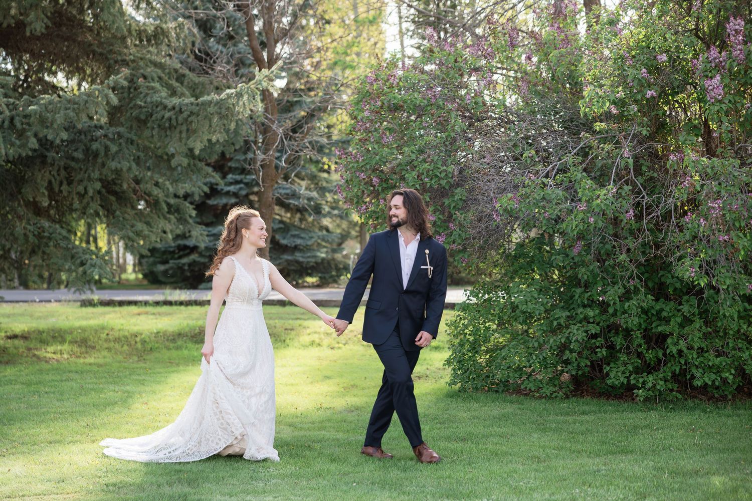 Bride and groom holding hands walking through lush green garden with pine and flowering trees on wedding day.