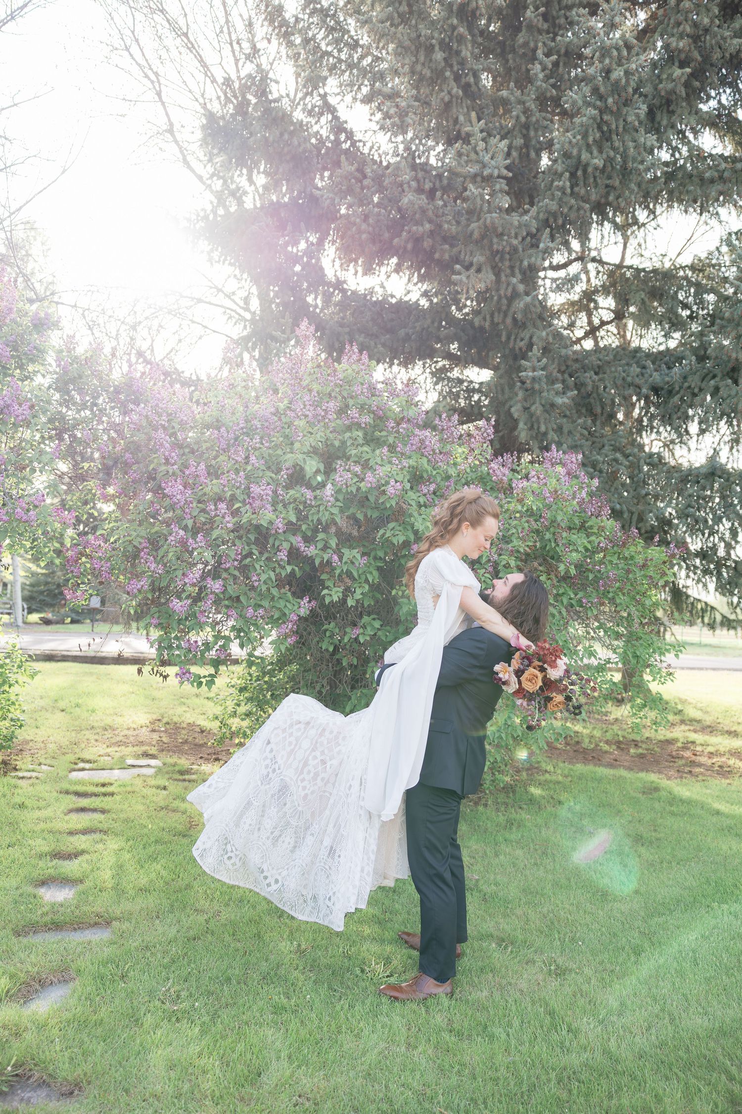 Groom lifts bride in white lace gown near blooming lilac bushes in sunny garden setting.