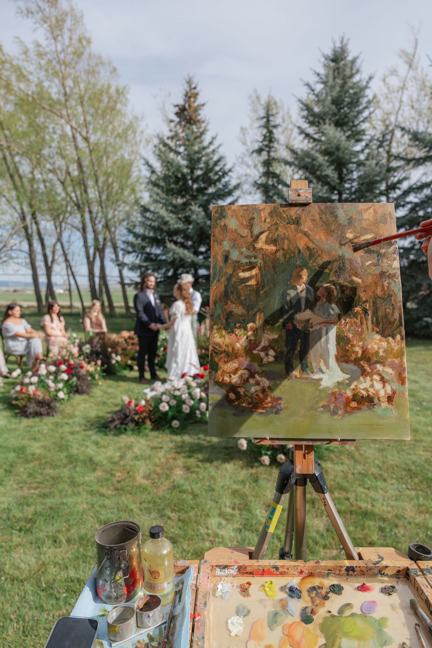 Artist painting wedding ceremony on easel outdoors with bride and groom exchanging vows in background.