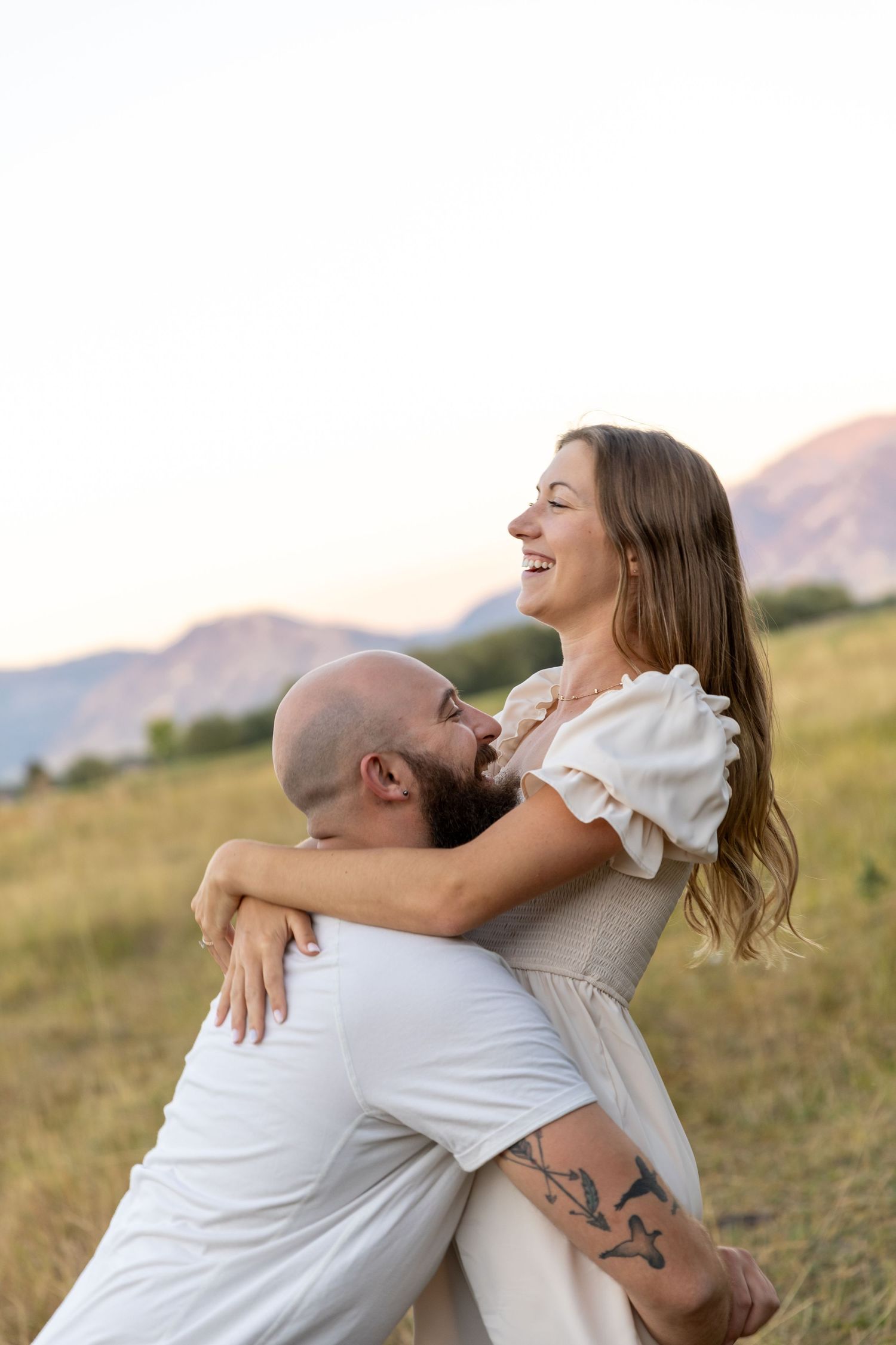A joyful couple embraces in a golden field with mountains behind them, the woman laughing as the man lifts her.