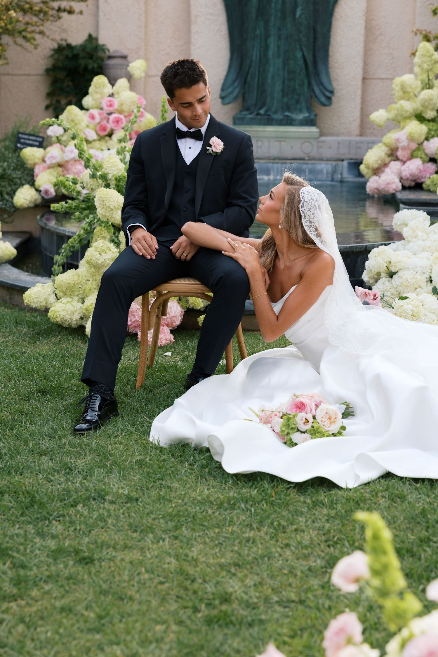 Bride in white gown sits on grass beside groom in tuxedo, surrounded by lush pink and white floral arrangements outdoors.