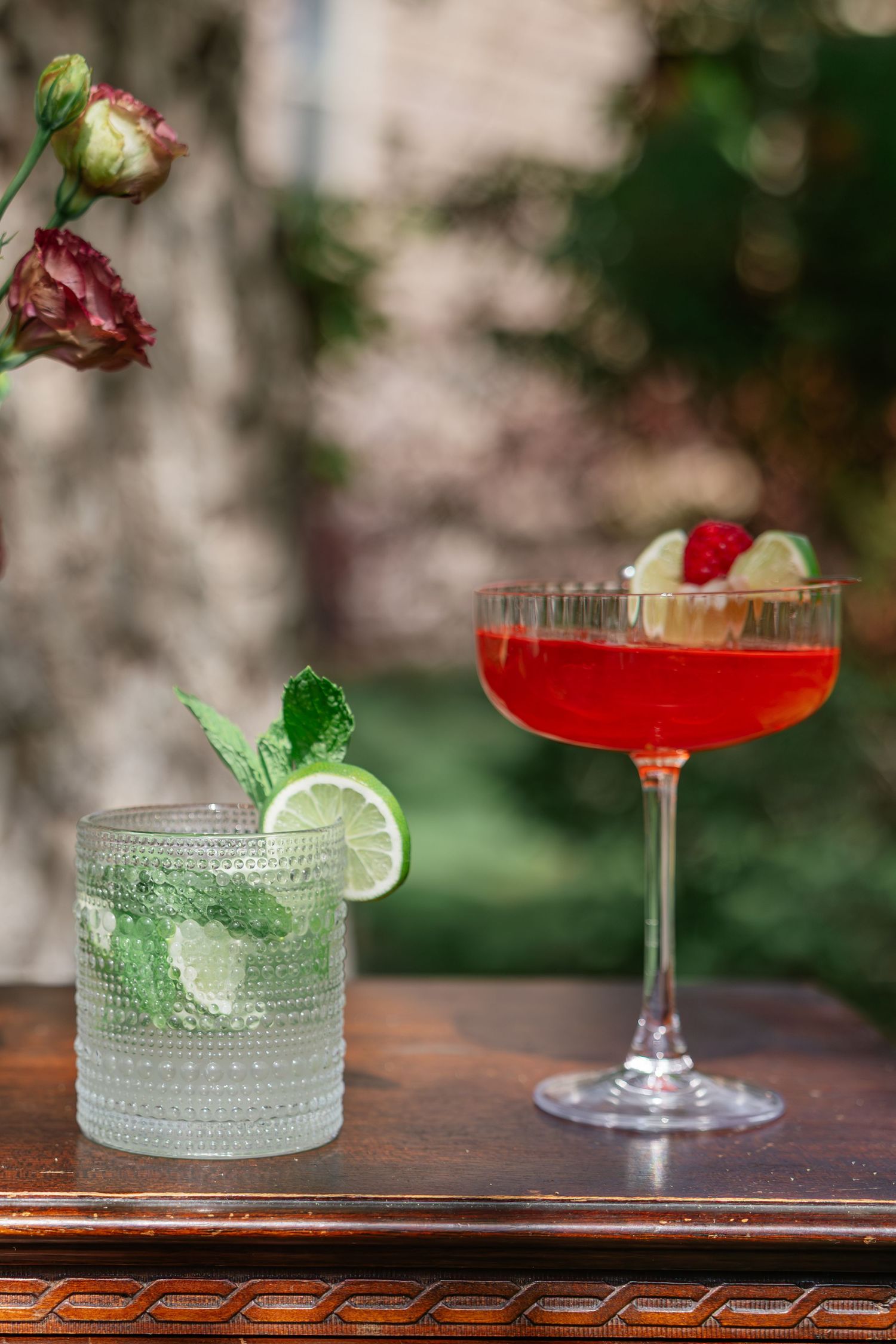 Two elegant wedding cocktails on a wooden table, featuring a mojito with lime and mint and a red raspberry coupe glass.