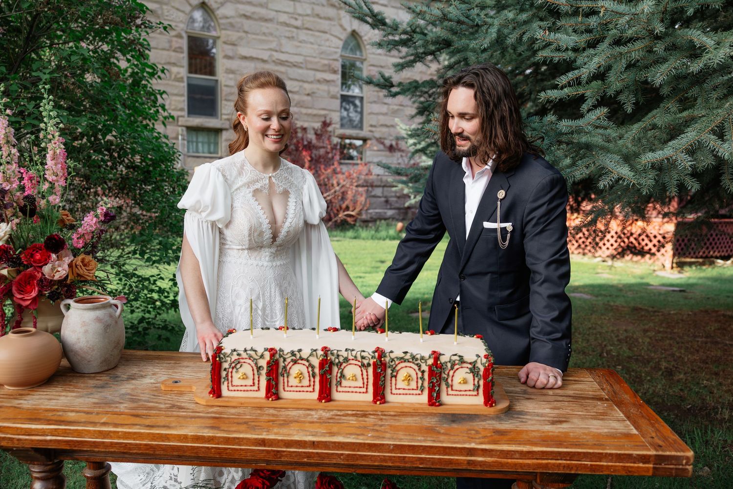 Bride and groom cut decorative wedding cake together on wooden table in outdoor garden setting.
