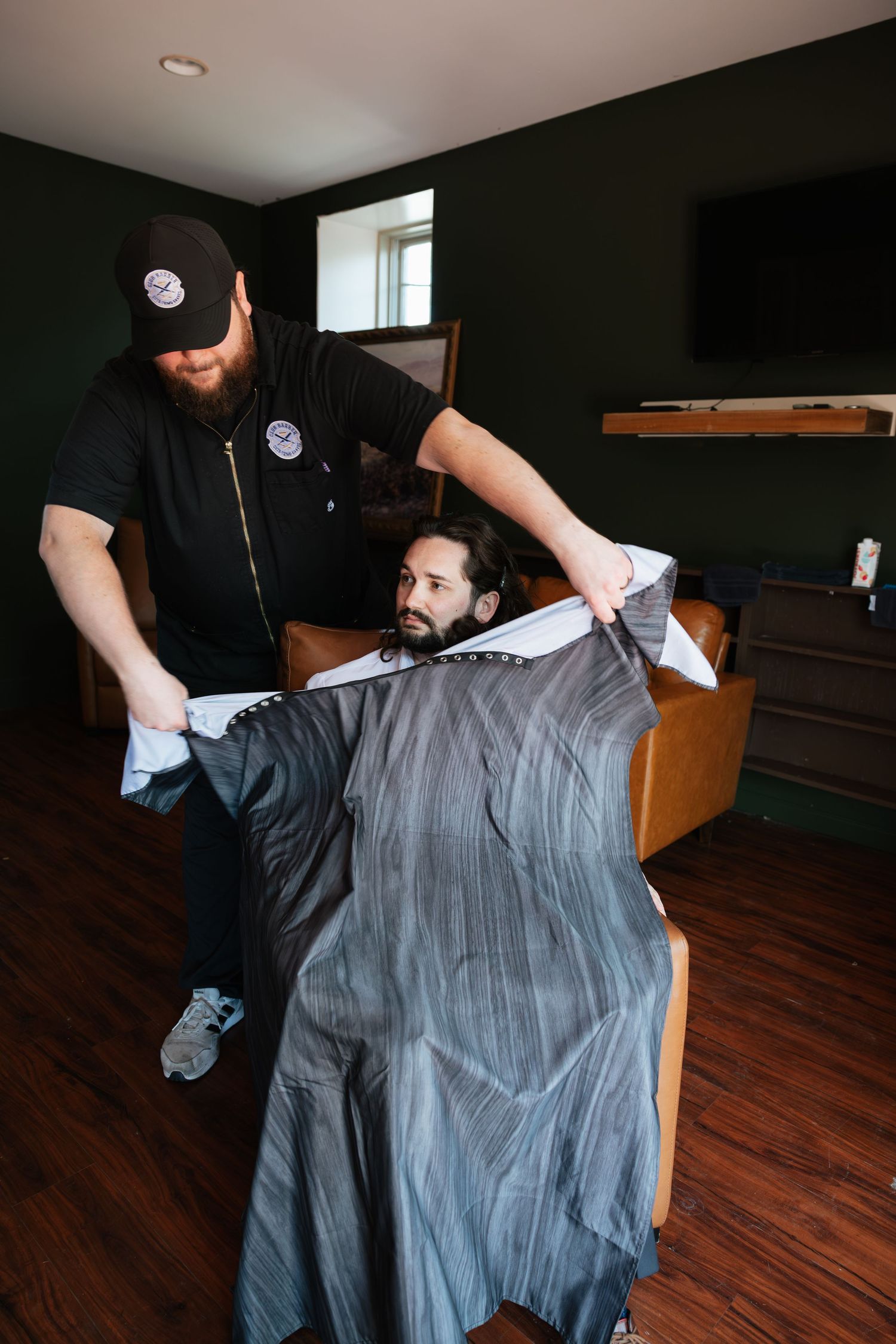 A barber drapes a gray cape over a bearded man seated in a chair during a wedding day grooming session.