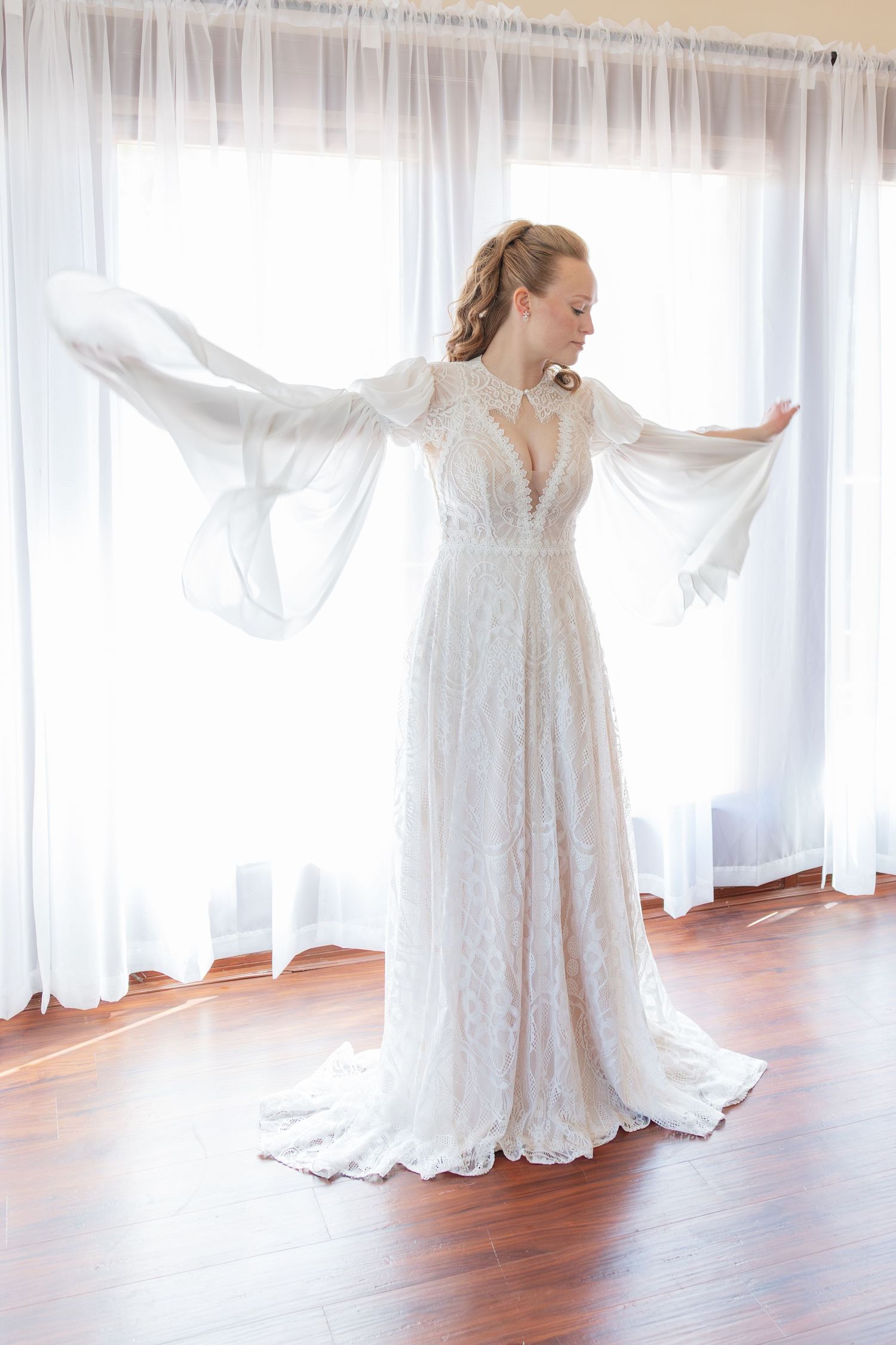 Bride in elegant white lace wedding gown with flowing bell sleeves standing by a bright window.
