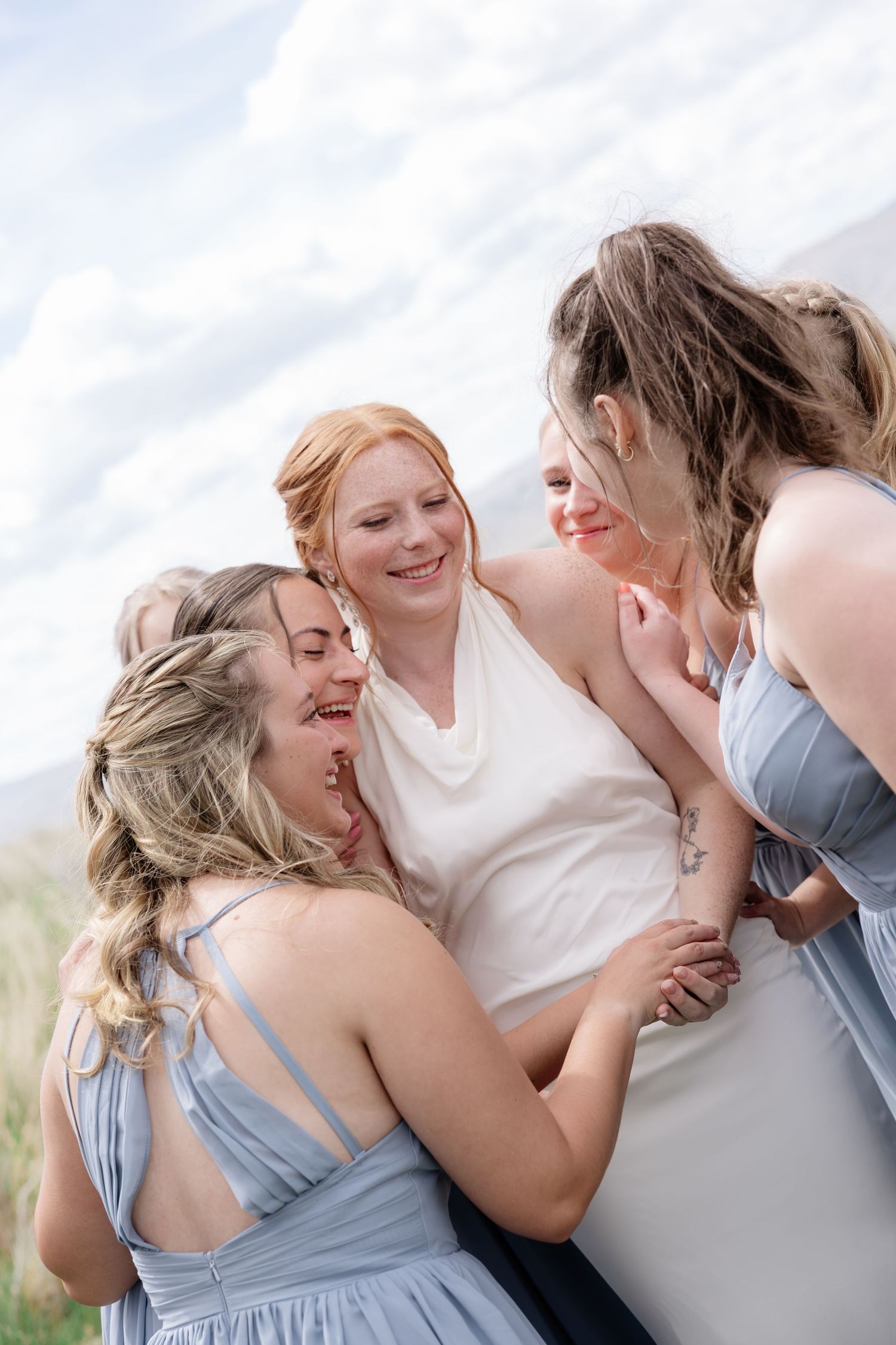 Bride in white dress laughing joyfully with bridesmaids in light blue gowns outdoors under a cloudy sky.