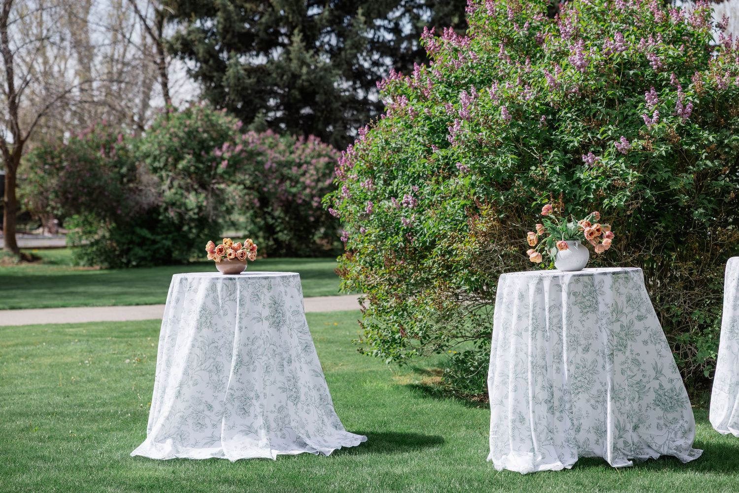 Outdoor wedding cocktail hour setup with lace-draped high-top tables and floral centerpieces on green lawn.
