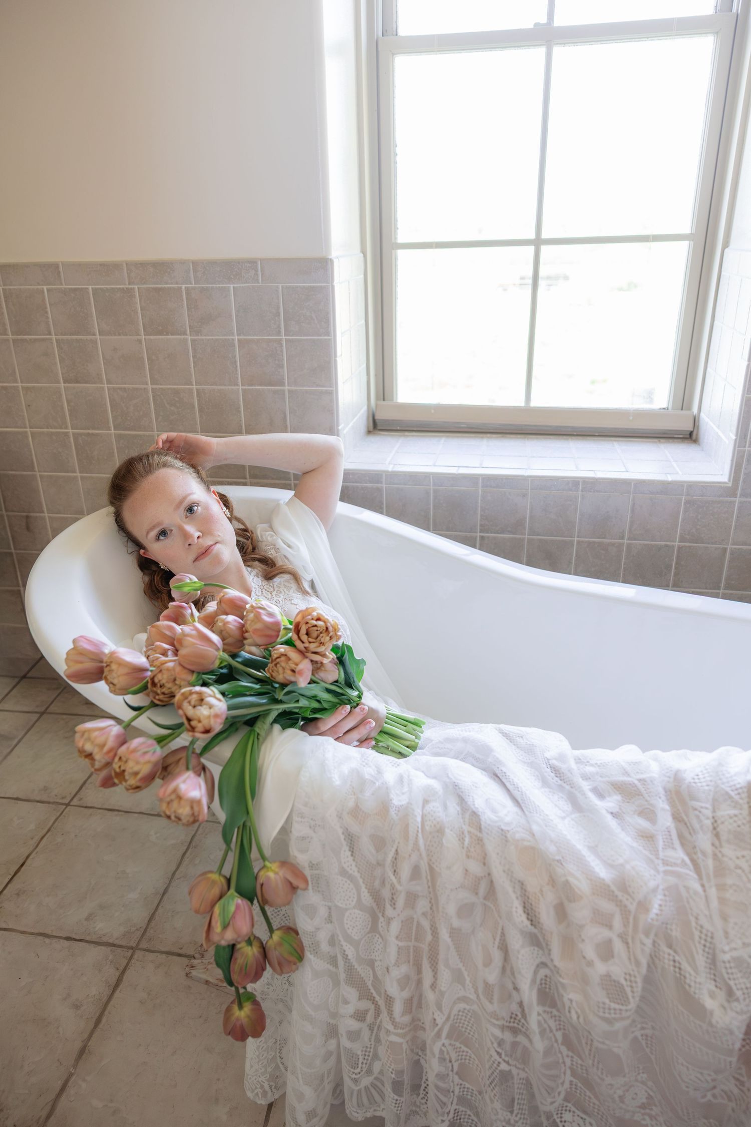 Bride in white lace dress relaxing in clawfoot bathtub holding bouquet of peach tulips near window.