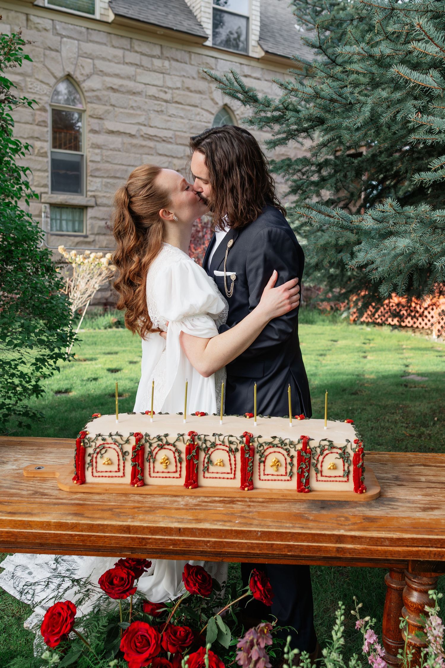 Bride and groom share a kiss behind an ornate rectangular wedding cake with red roses outdoors.