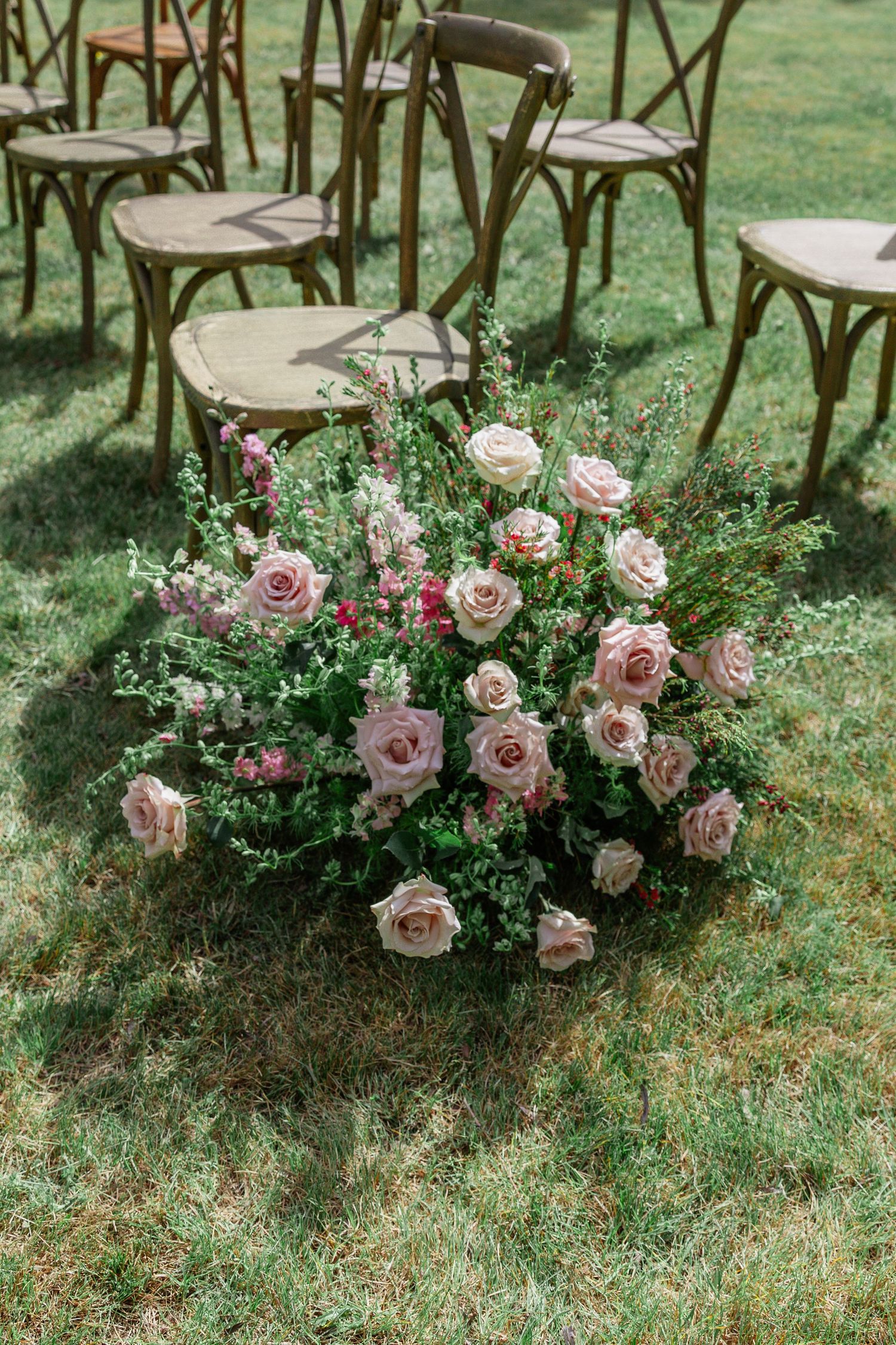 Lush pink roses and greenery floral arrangement on grass at an outdoor wedding ceremony with wooden chairs.