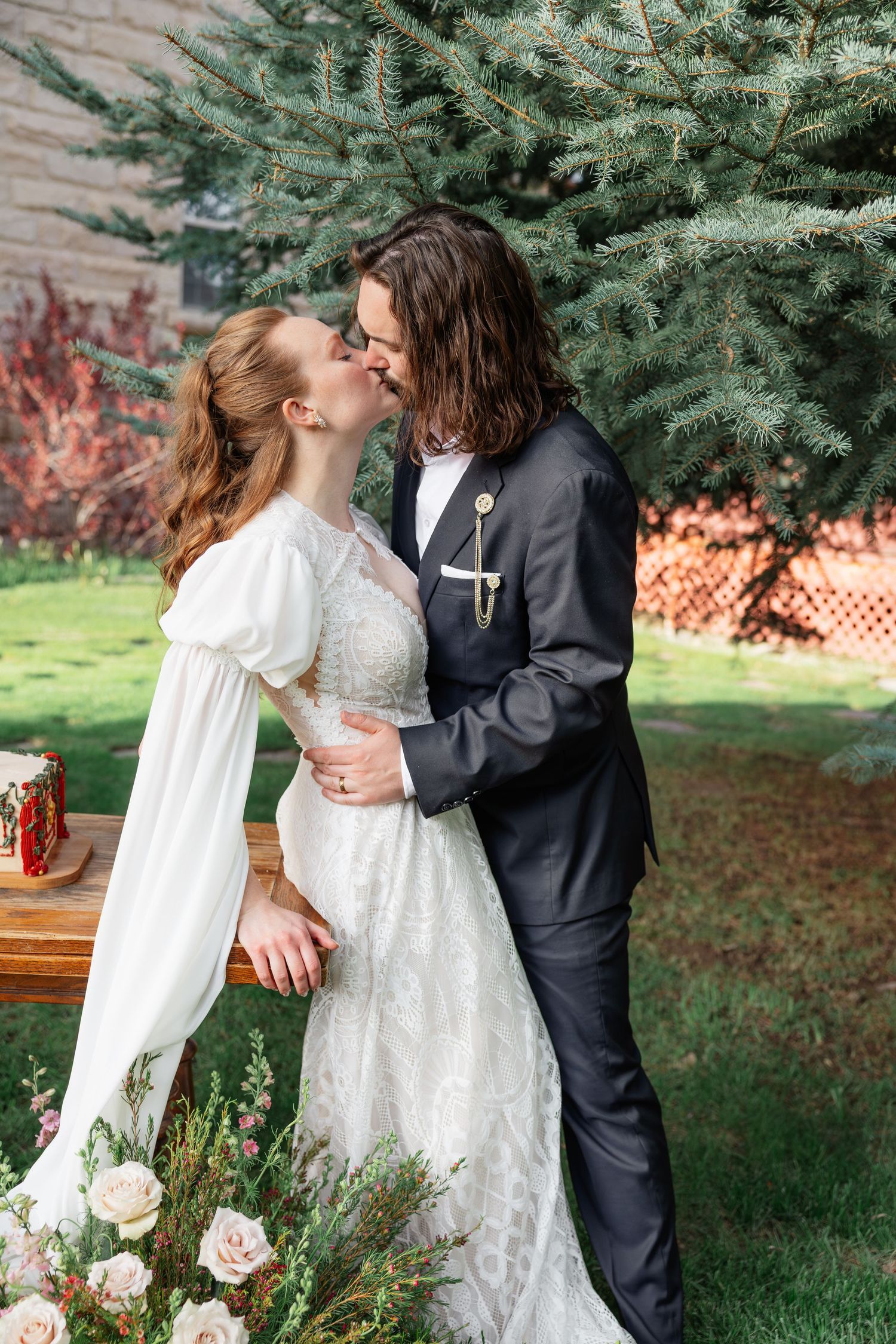 Bride and groom sharing romantic kiss outdoors near blue spruce tree, bride in lace gown, groom in dark suit.