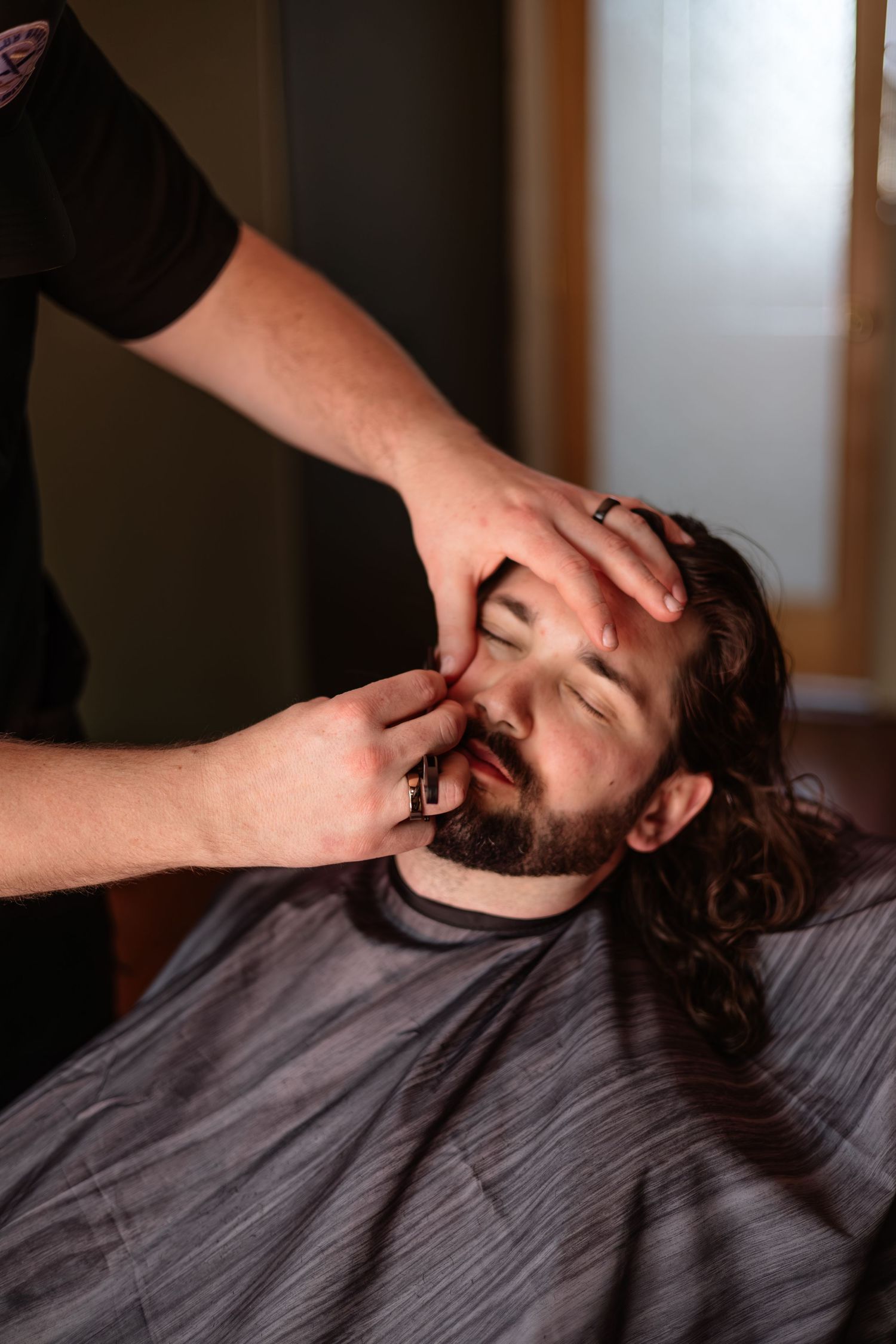 Barber grooming a bearded groom with long dark hair at a barbershop before his wedding day.