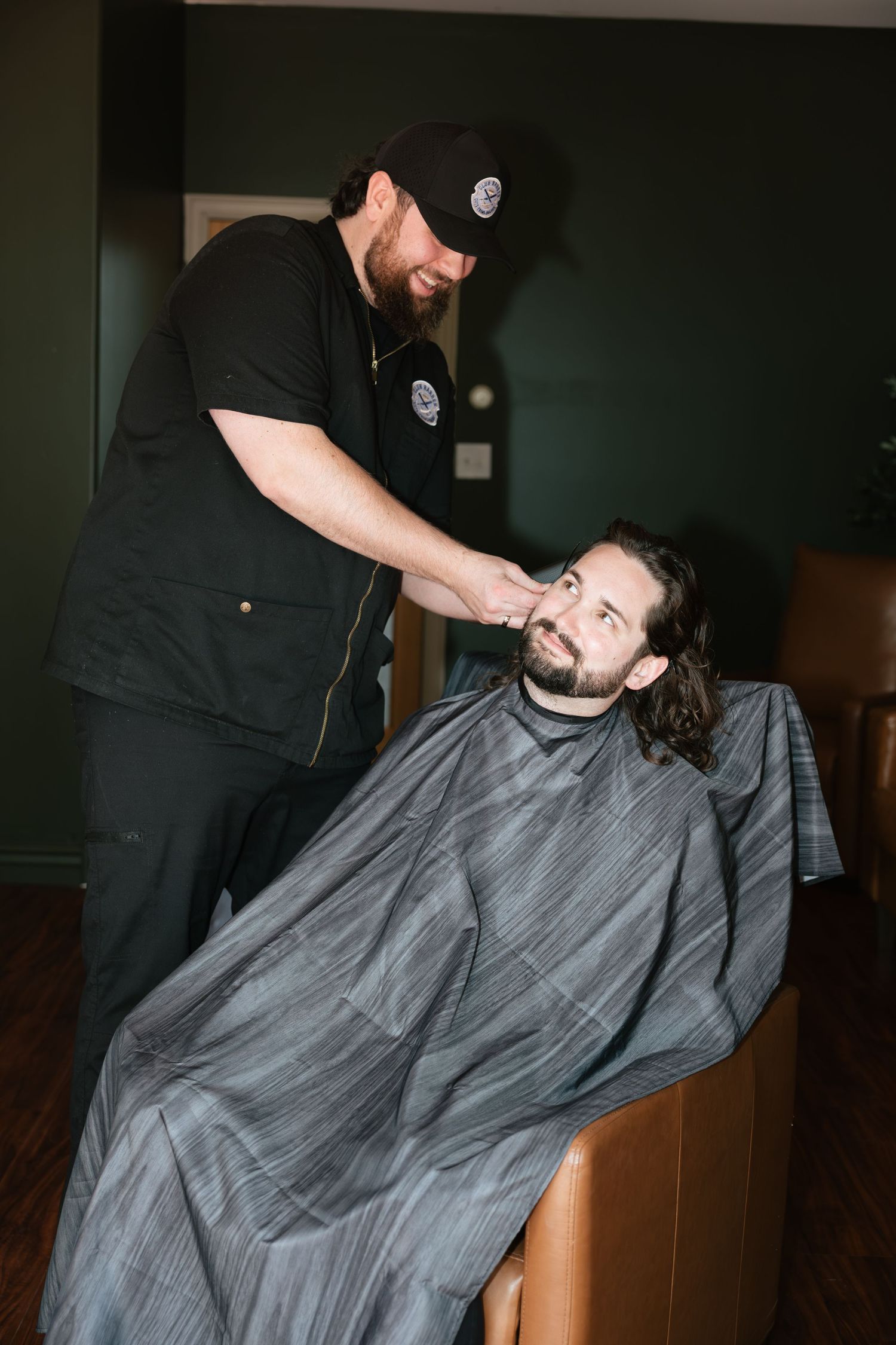 A smiling barber in black uniform gives a haircut to a bearded man in a gray cape at a barbershop.