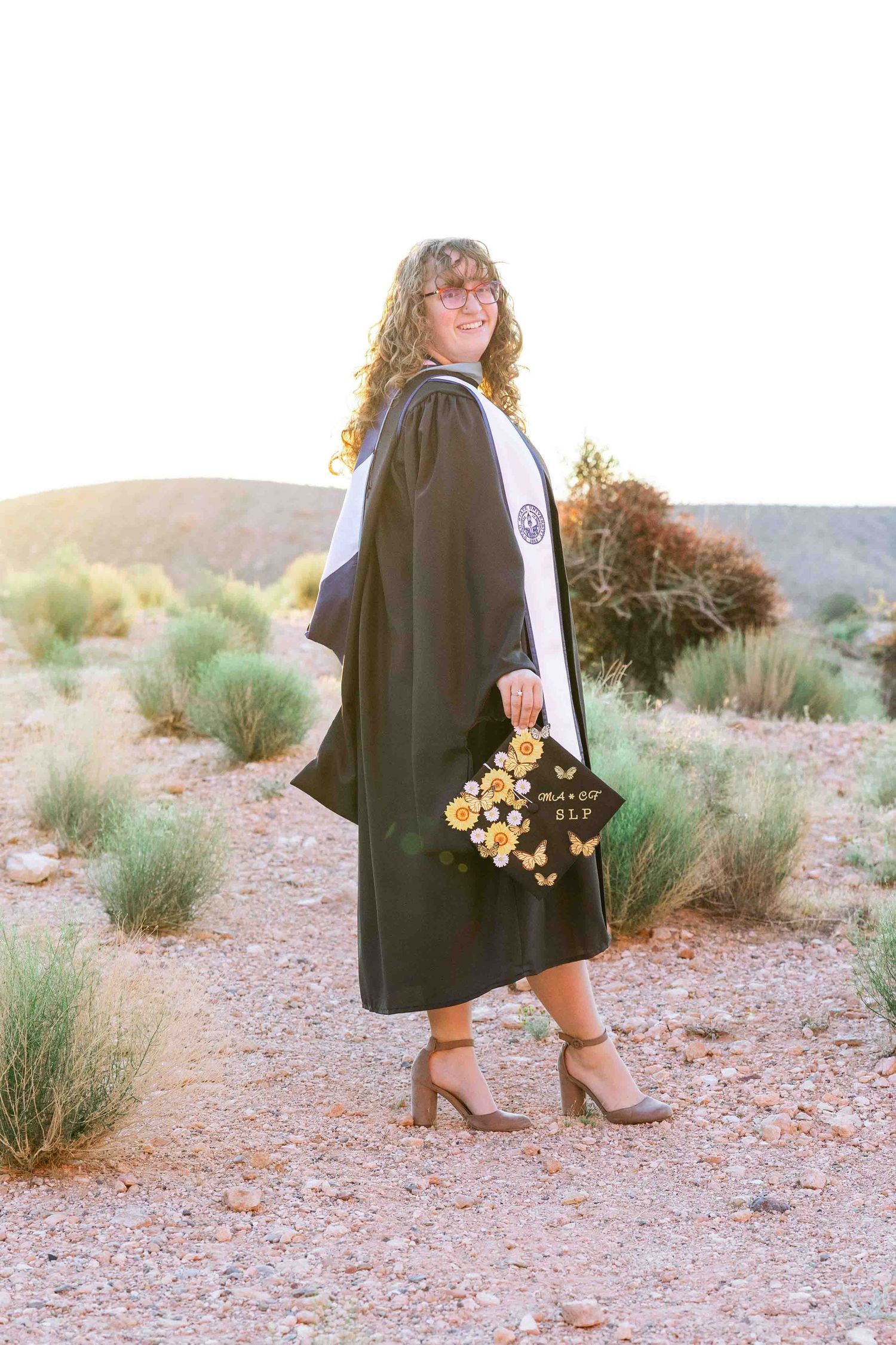 Woman in black graduation gown holding decorated cap with flowers poses outdoors in desert landscape at golden hour.