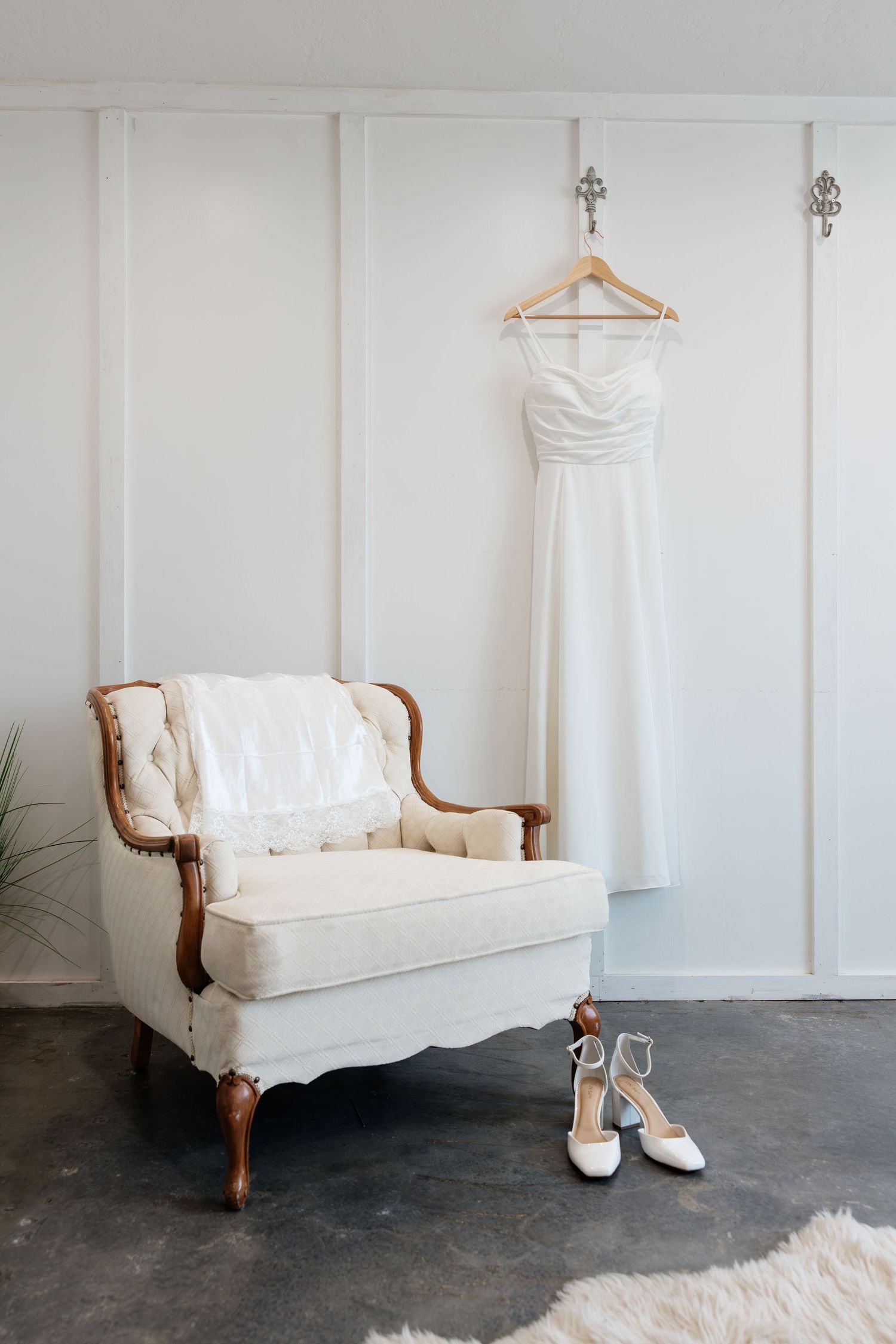 Elegant white wedding dress hanging on wall hook beside vintage armchair with bridal heels on concrete floor.