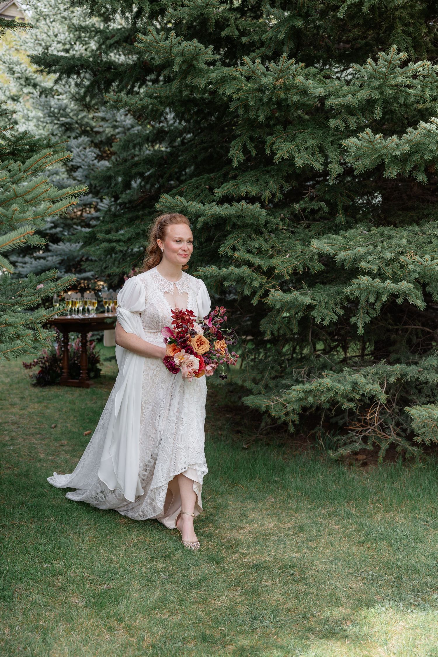 Smiling bride in white lace wedding dress carries colorful bouquet while walking on green lawn.