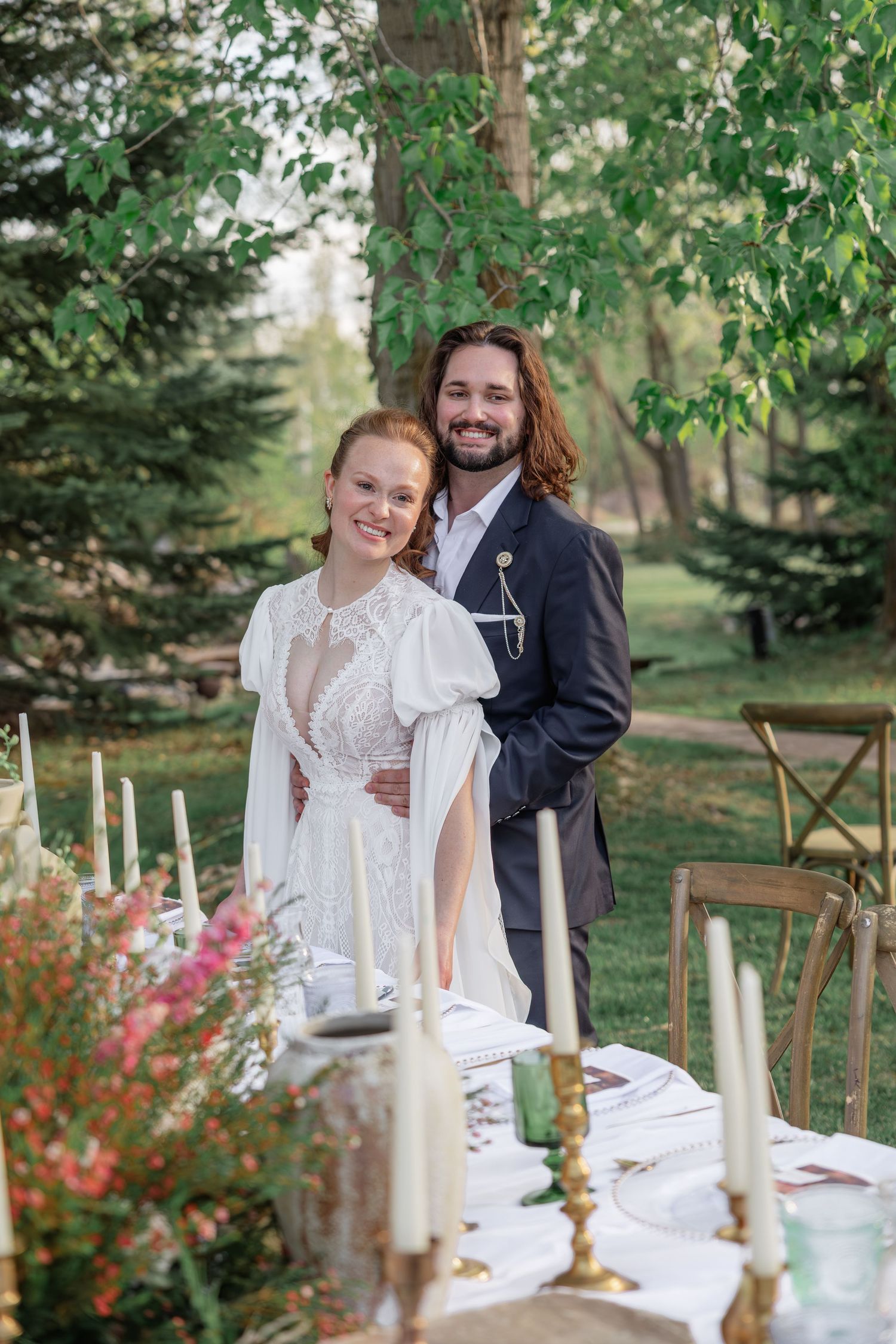 Smiling bride and groom posing at outdoor reception table with candles and floral arrangements.