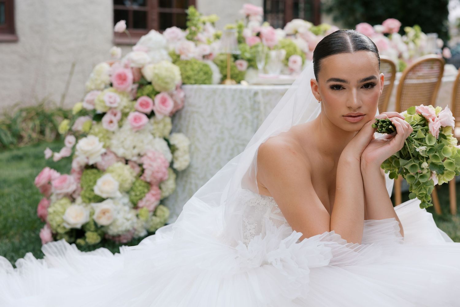 Elegant bride in a white tulle gown with veil poses near lush pink and green floral arrangements outdoors.