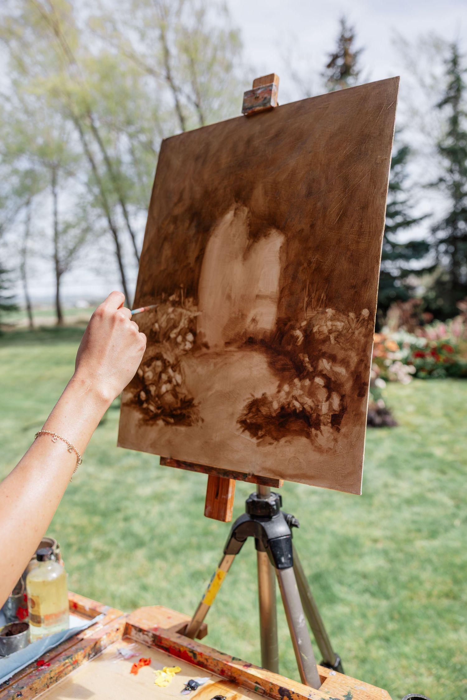 Artist painting a brown monochromatic landscape on canvas outdoors on an easel in a lush green garden.