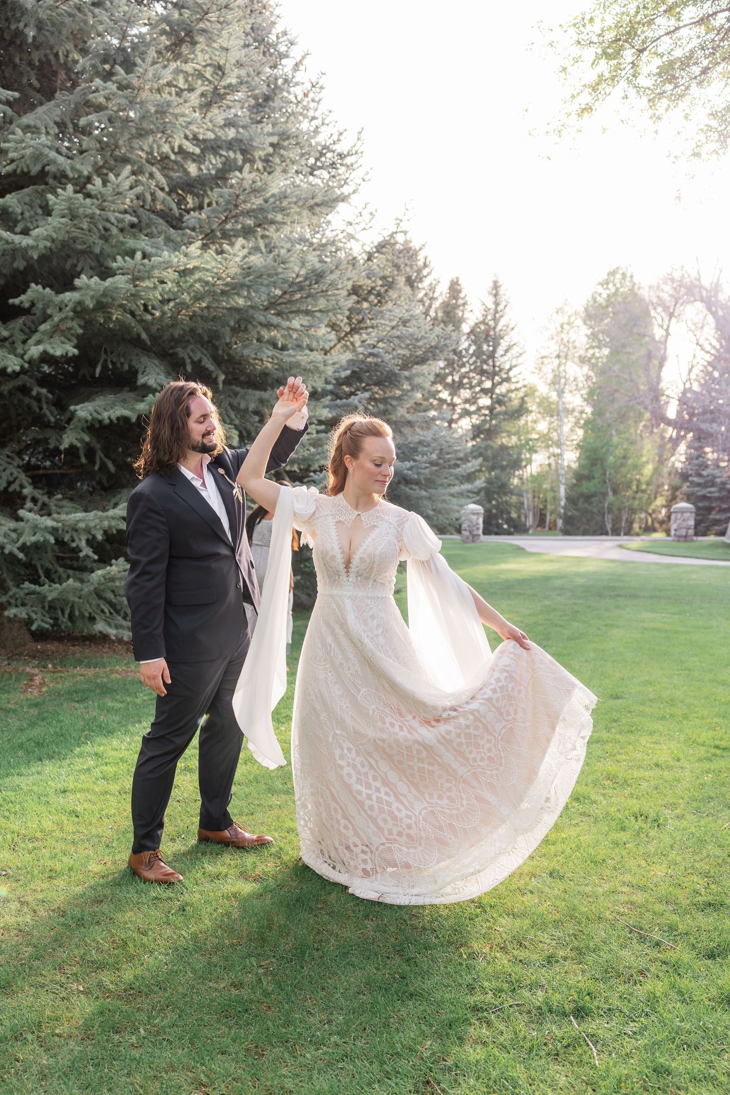 Bride and groom dancing outdoors in garden, groom spinning bride in lace wedding dress.