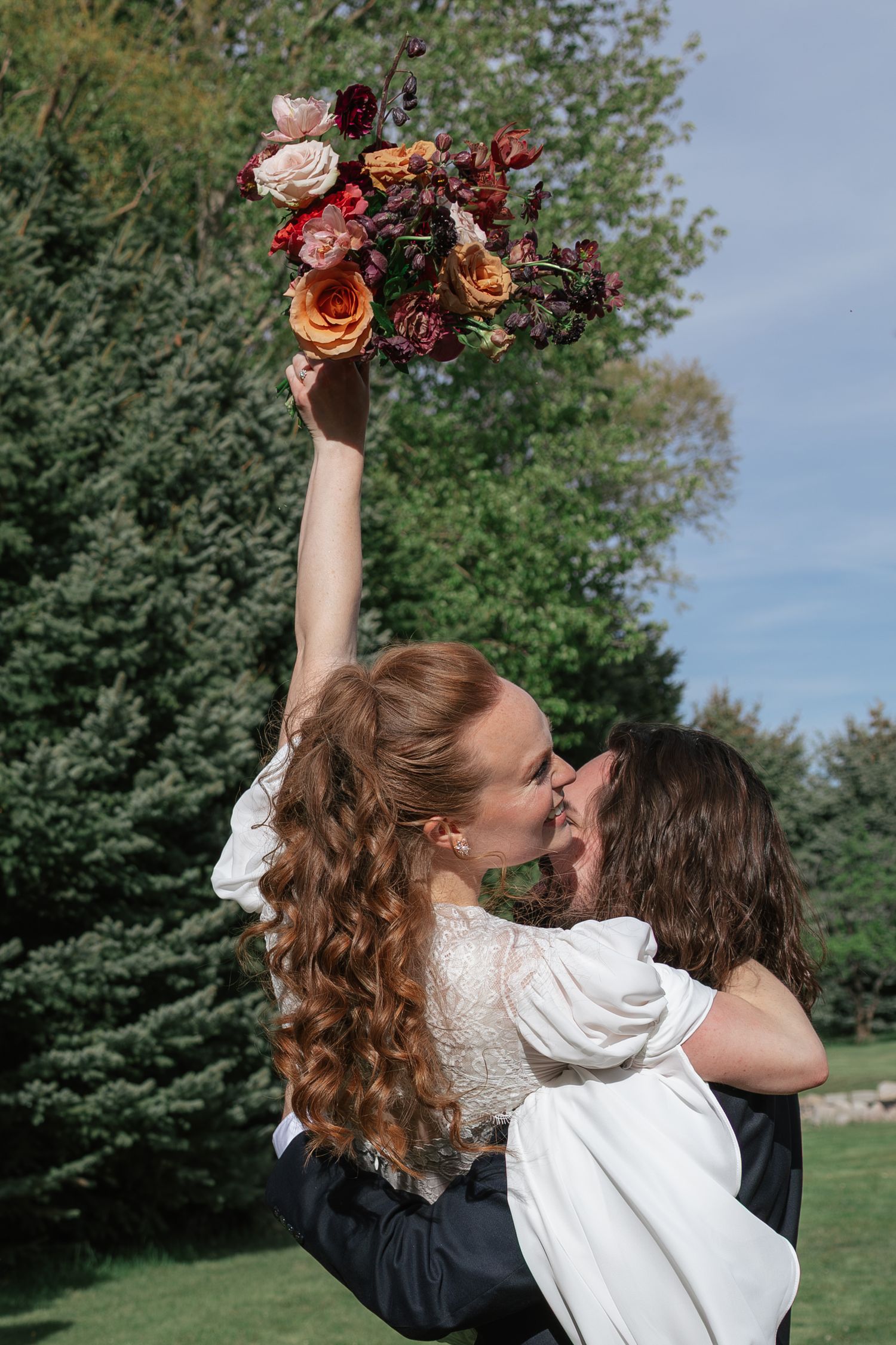 Groom lifting bride while kissing as she raises bouquet of red and orange roses against backdrop of green trees.
