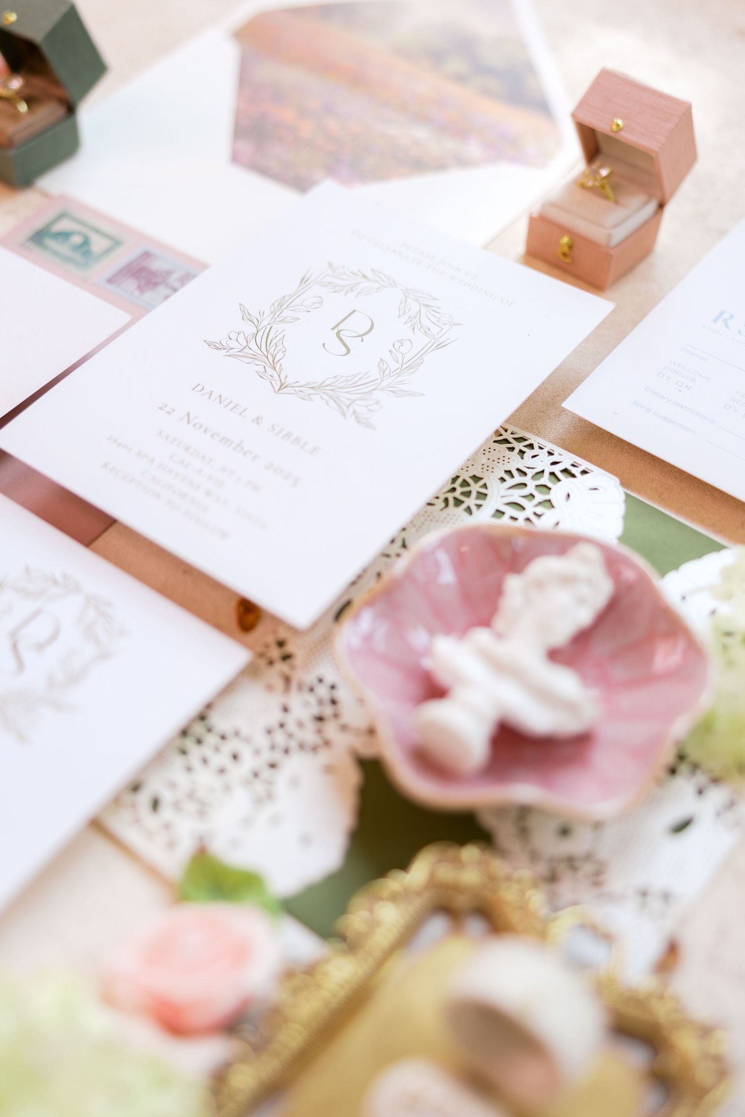 Elegant wedding stationery suite with gold monogram crest, pink velvet ring box, and vintage decorative accents on a styled flat lay.
