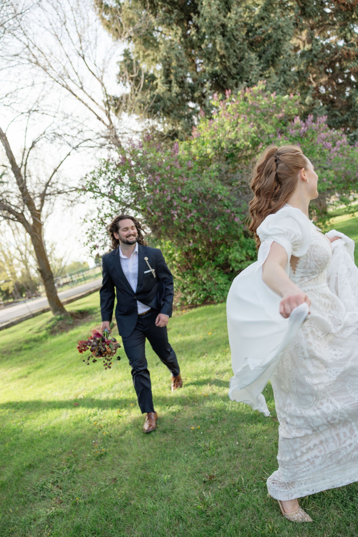 Groom walking toward bride in white lace dress on green lawn with spring trees behind.
