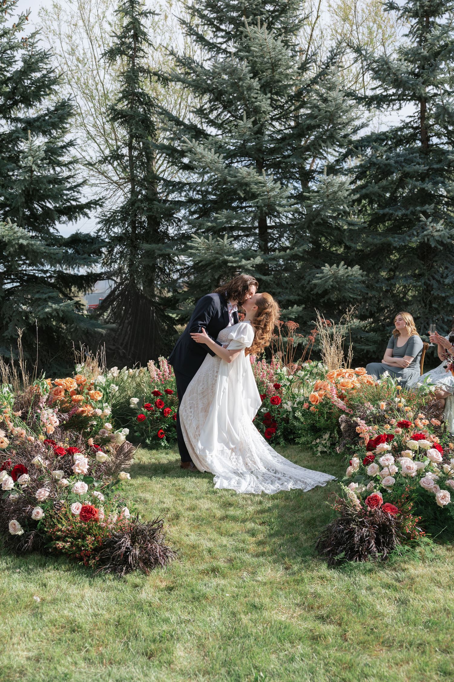 Bride and groom sharing first kiss at outdoor wedding ceremony surrounded by lush colorful floral arrangements and evergreen trees.