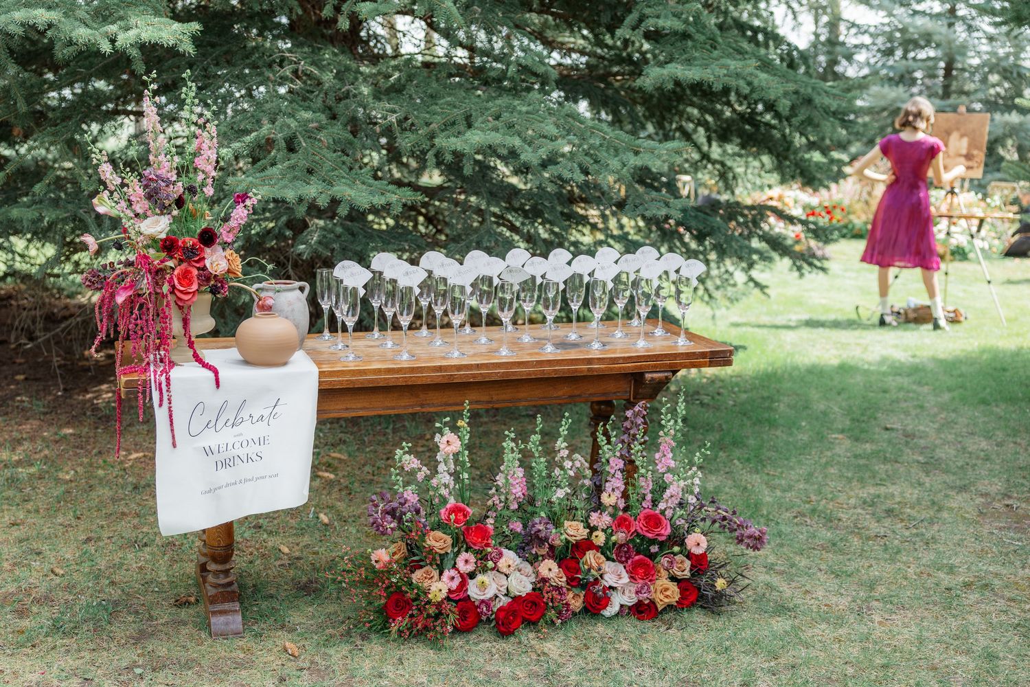 Outdoor wedding welcome drink station with floral arrangements, champagne flutes on wooden table, and colorful garden blooms.