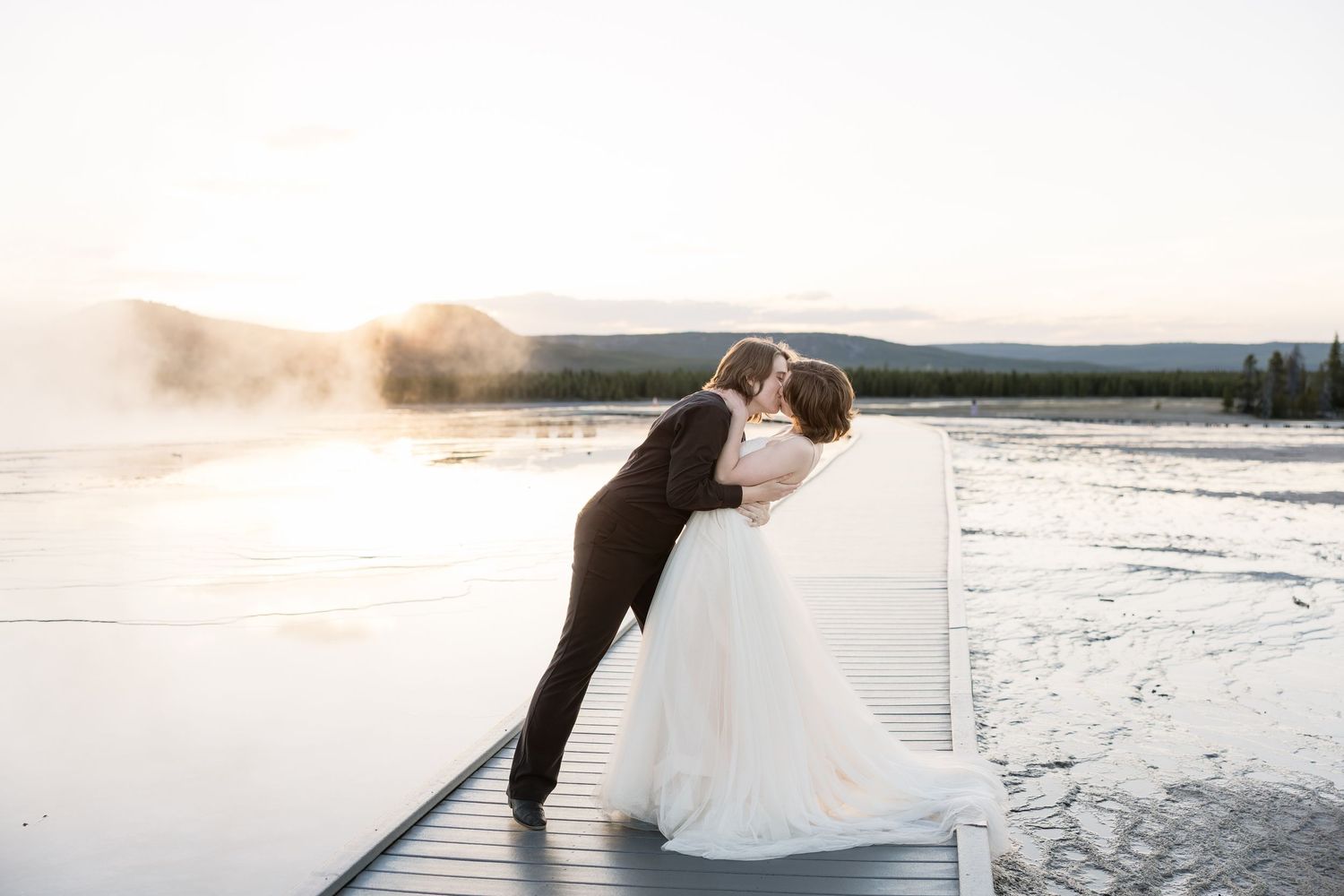 Bride and groom share a romantic dip kiss on a dock at sunset with misty lake and mountains in background.