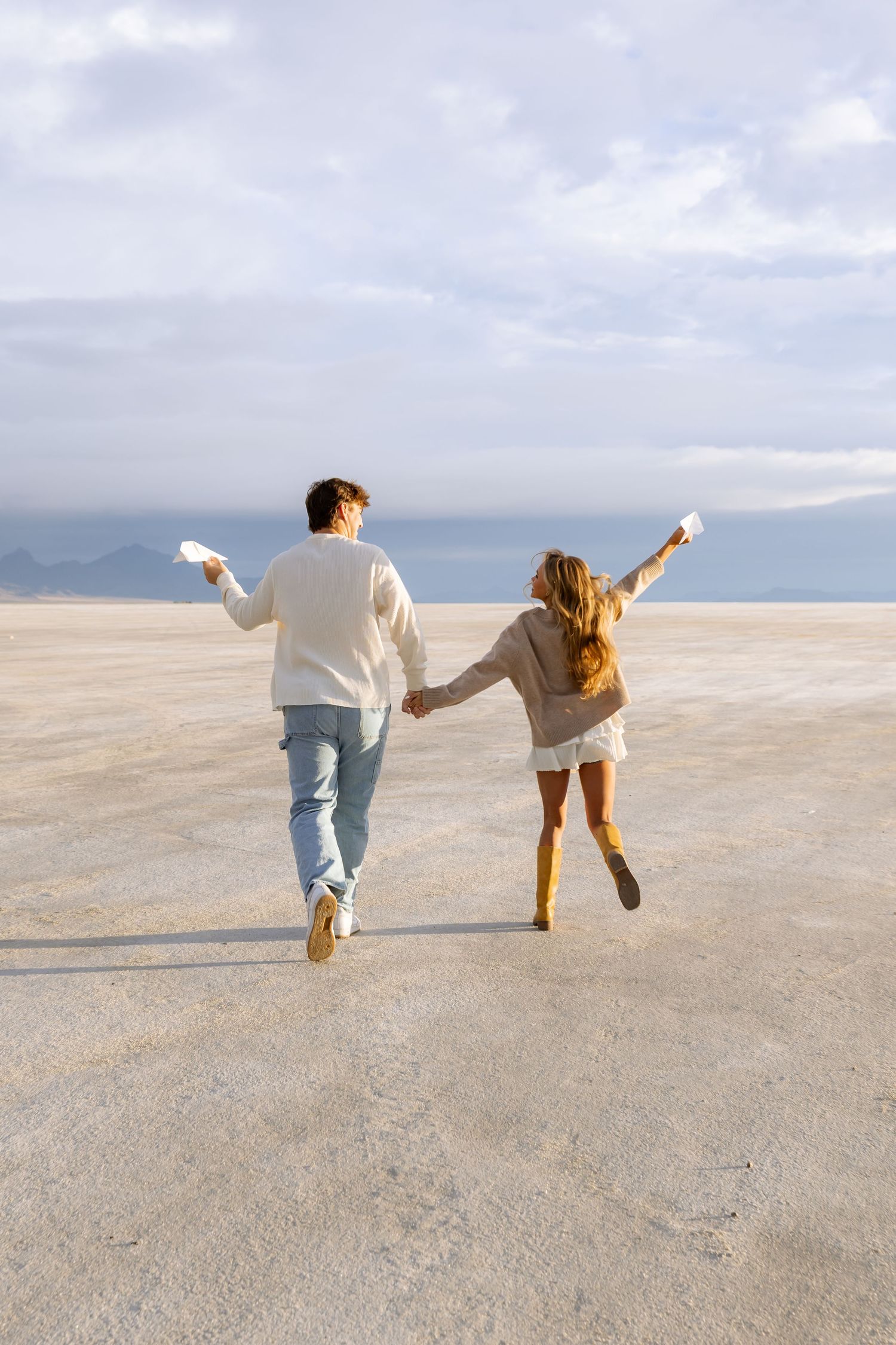 Couple running playfully on a salt flat under cloudy skies.