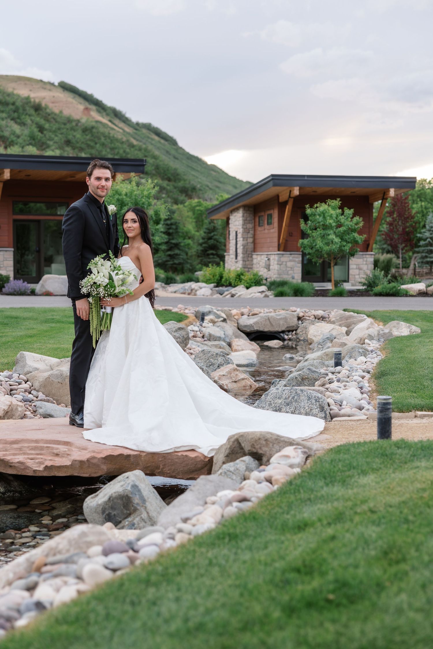 Bride and groom pose on stone bridge surrounded by lush greenery and mountain backdrop at elegant outdoor wedding venue.