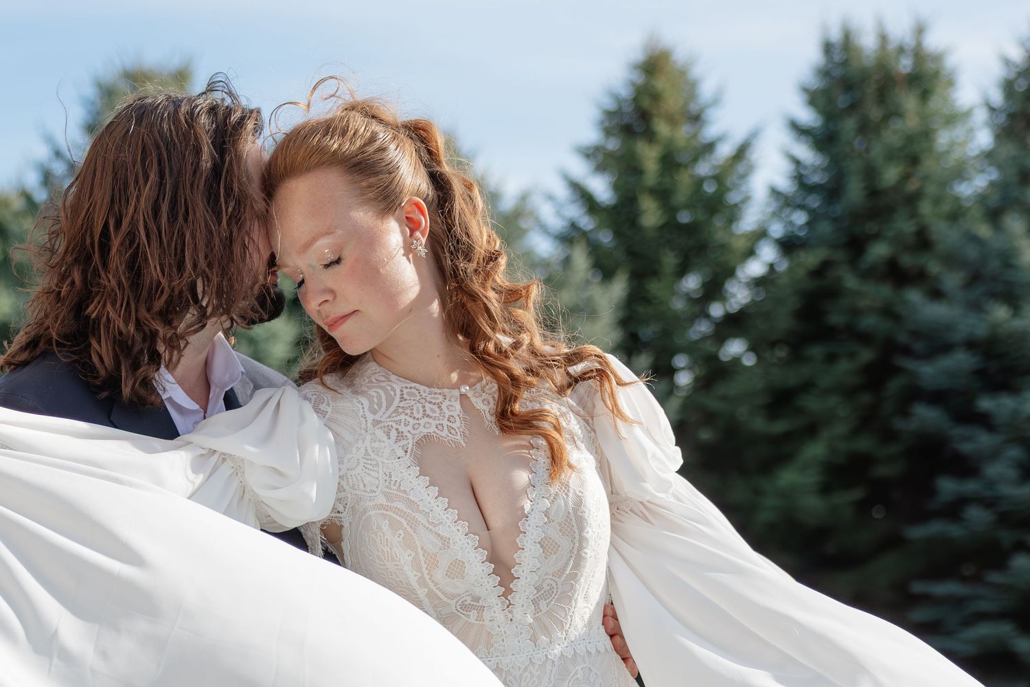 Romantic close-up of bride in lace wedding gown and groom sharing an intimate moment outdoors with pine trees behind them.
