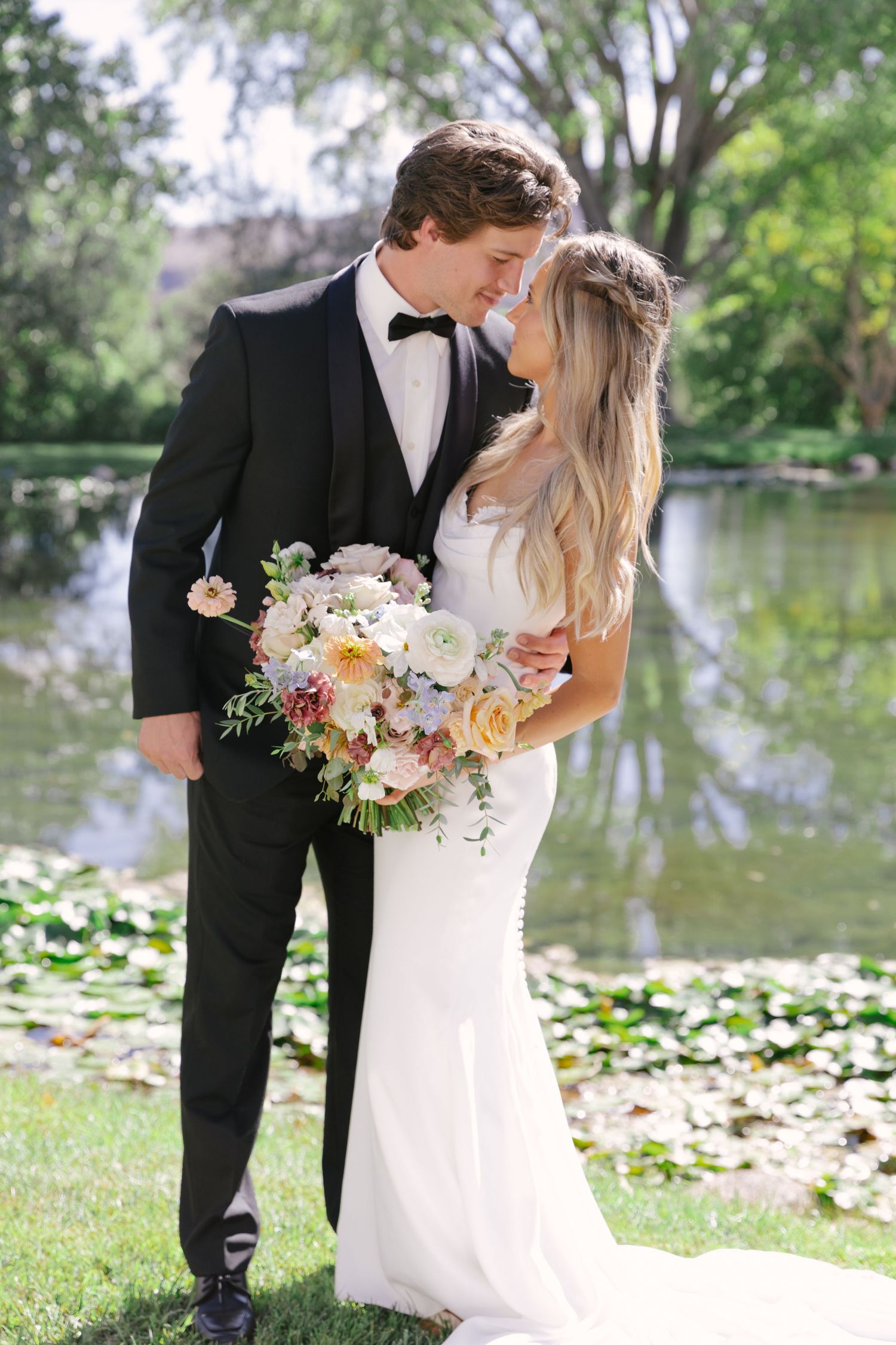 Bride and groom share a romantic moment by a lily pond, she holds a pastel bouquet, he wears a black tuxedo.