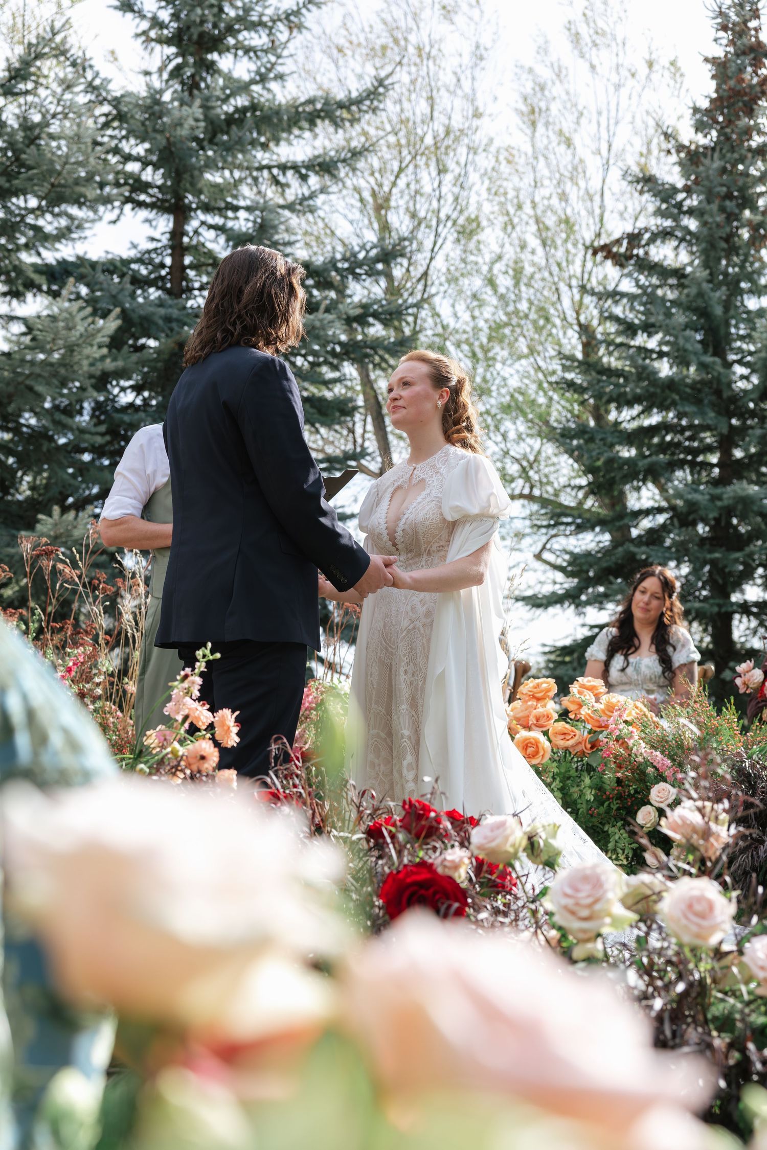 Bride and groom exchange vows outdoors surrounded by lush floral arrangements and evergreen trees at a garden wedding.
